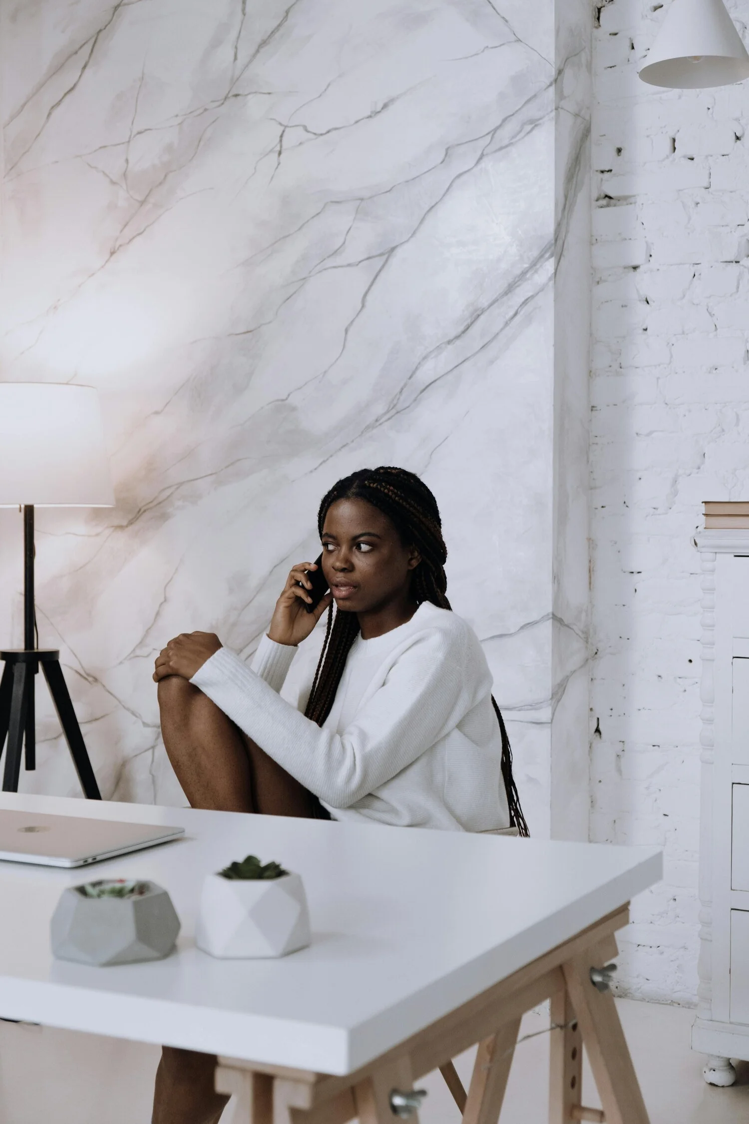 A woman with dark braided hair sitting at a white desk, holding a phone to her ear, in a modern interior with white marble and brick wall, a lamp, and small potted plants.
