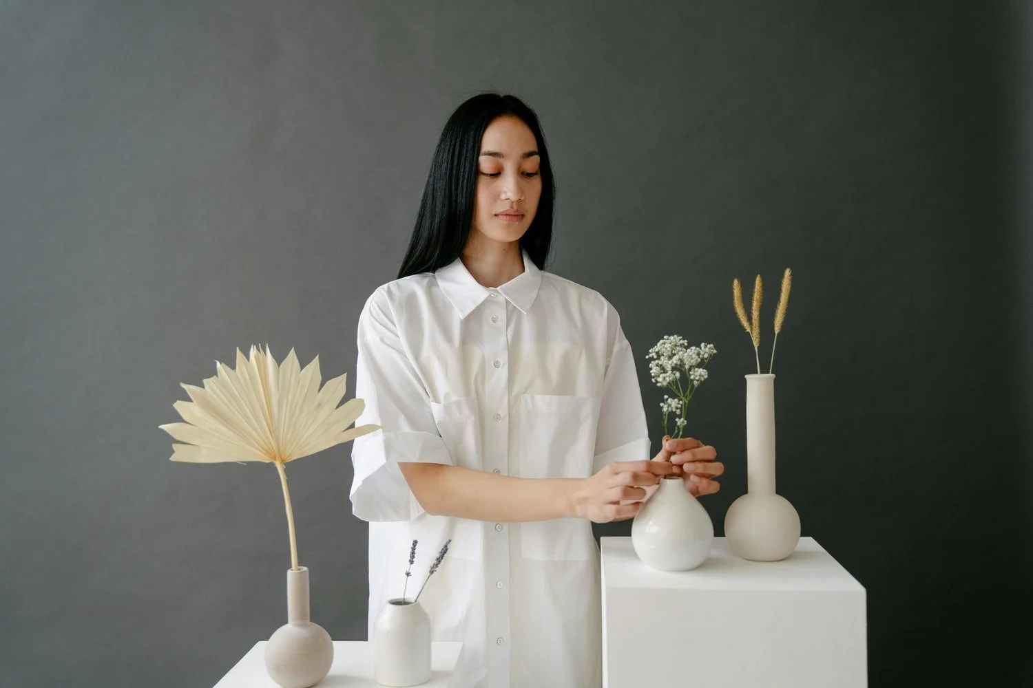 A woman with long black hair wearing a white shirt arranging flowers in vases on white pedestals against a dark gray background.