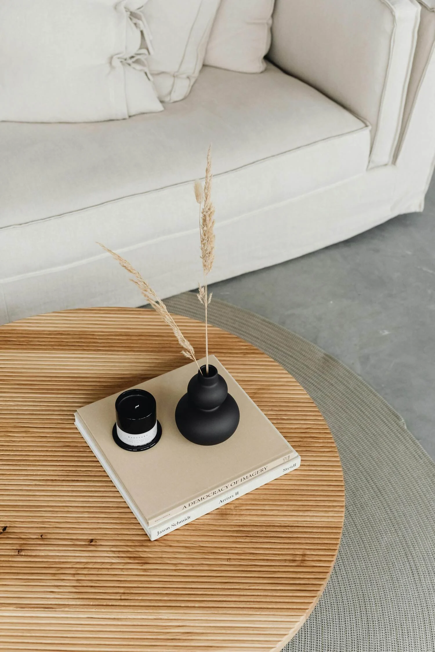 A round wooden coffee table with a beige book, a black vase with dried grass, and a small black container on top, situated in front of a white sofa with cushions.
