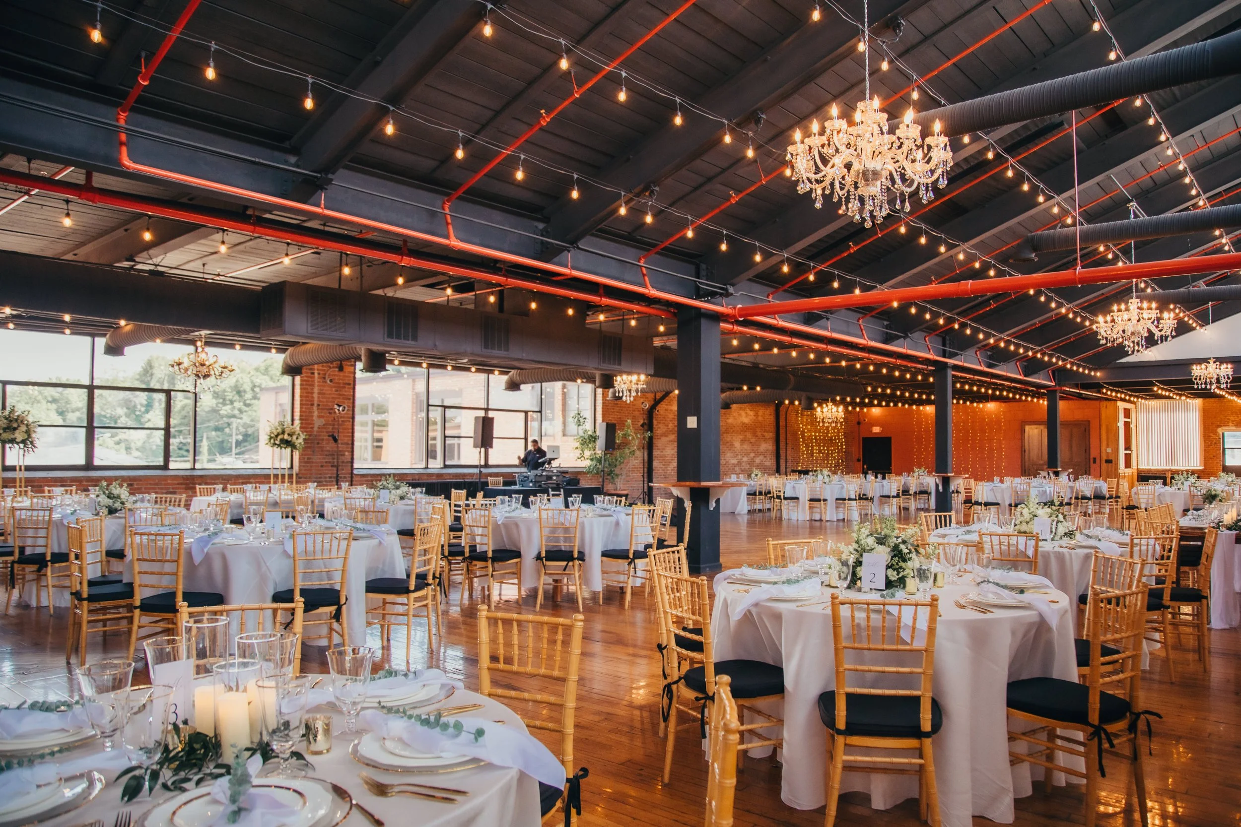 Elegant event hall with round tables decorated with white tablecloths, floral centerpieces, candles, and place settings. Gold chairs with black cushions fill the room, which has large windows, exposed brick walls, and string lights hanging from the ceiling. Chandeliers hang from the ceiling, and a DJ is set up near the windows.