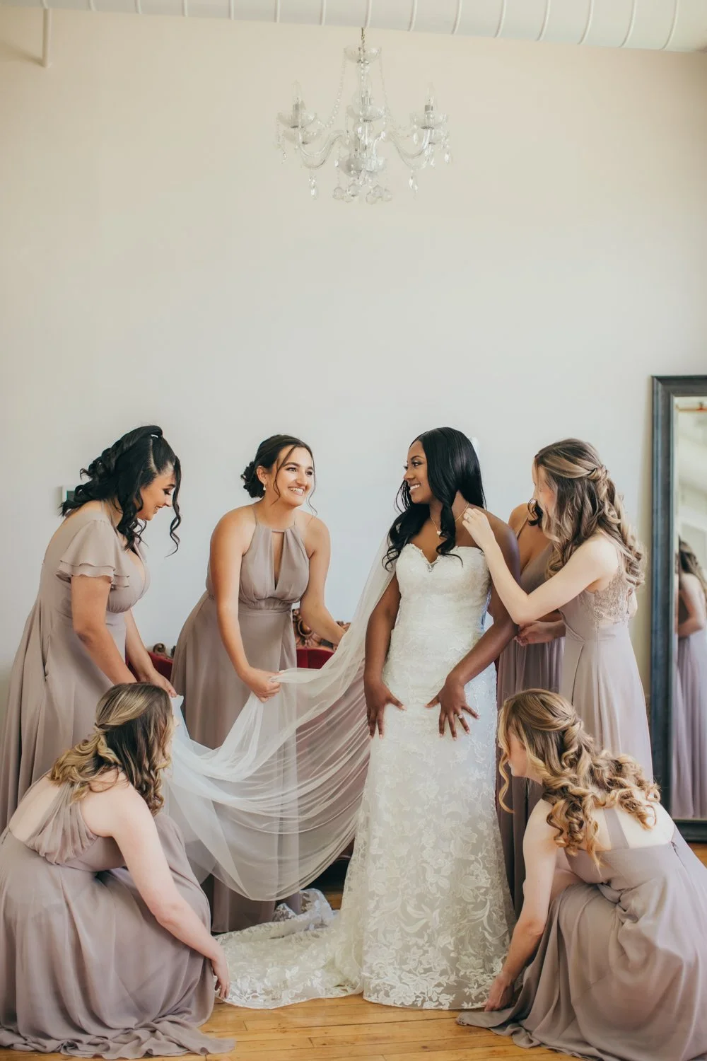 A bride with black hair in a white lace wedding dress is surrounded by her bridesmaids who are helping her with her veil and dress in a room with a chandelier and a large mirror.