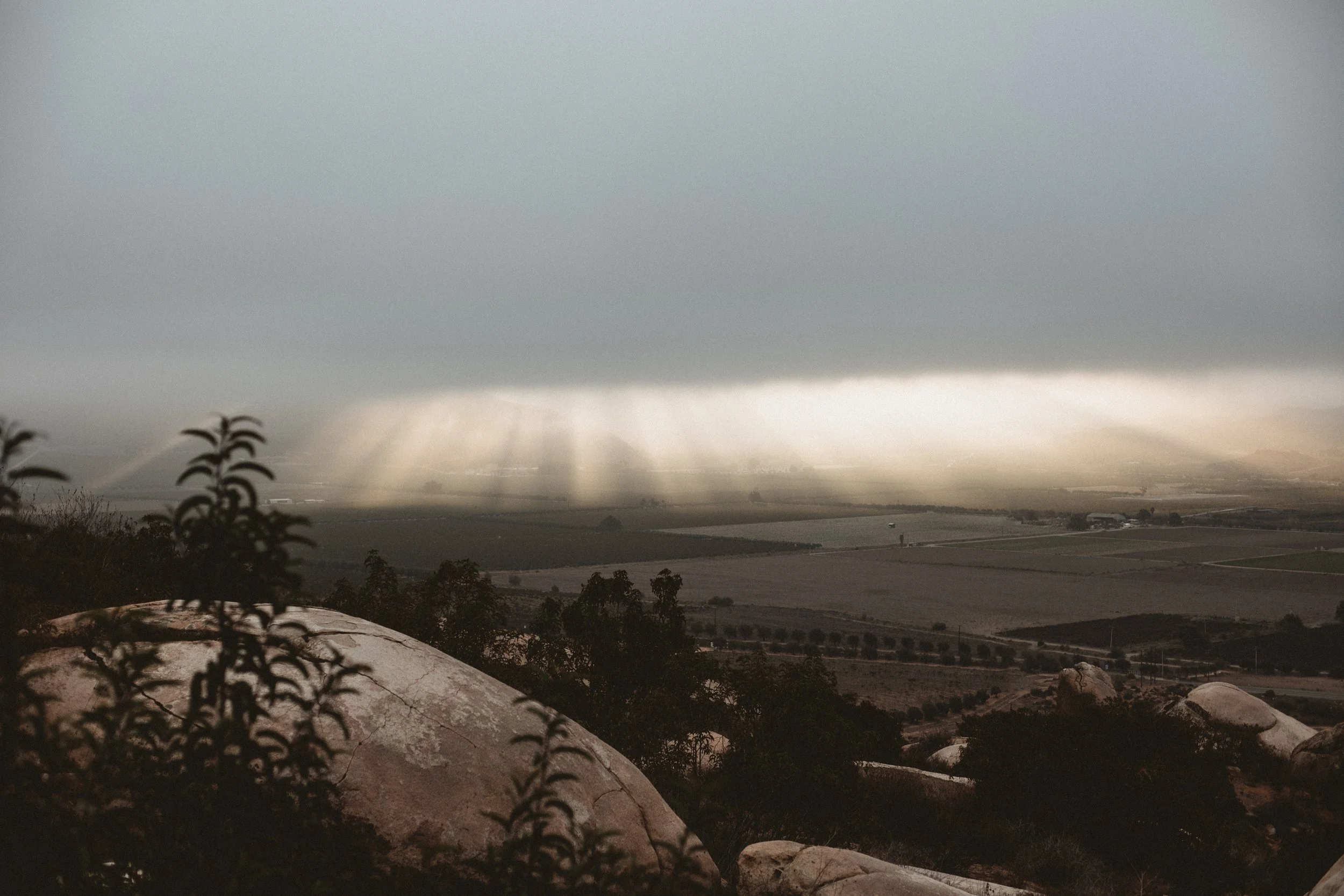 View of valley below large boulder and sun rays outpouring from clouds