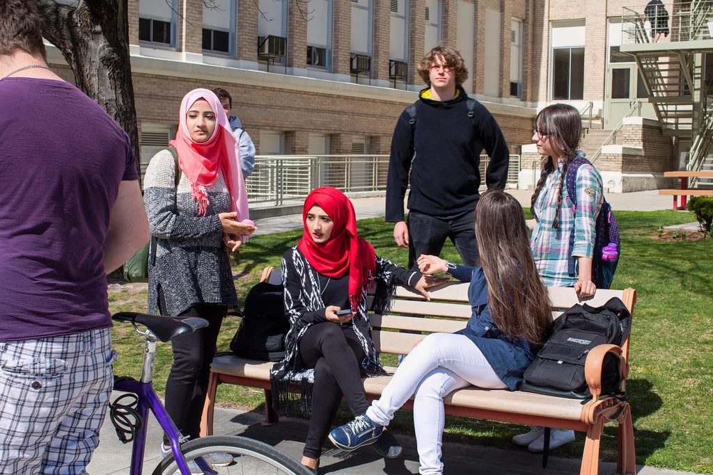 Zahraa Naser, 18, center, in red hijab, at Boise High School with her sister, standing, left, and best friend (in blue on the bench), who is, like Zahraa, from Iraq, image by Angie Smith, courtesy of the New York Times, September 2016.