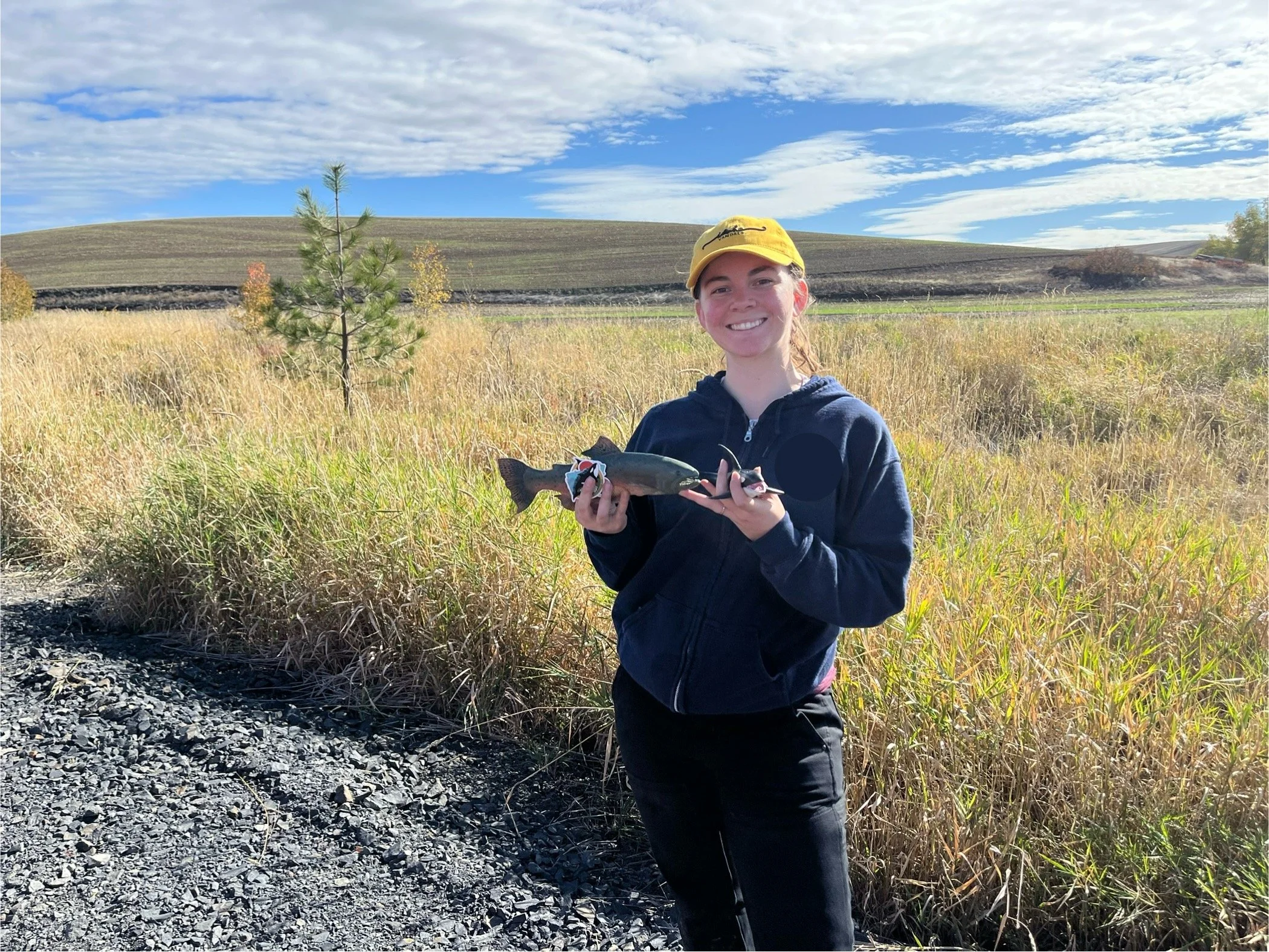 Erin is standing on a gravel roadside with pasture in the background.  She is holding a model of a salmon and an orca.