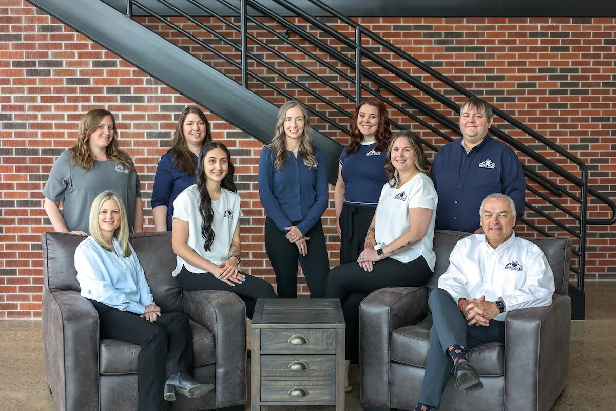 Group of nine people in business casual attire sitting and standing inside a building with a brick wall and a staircase behind them.