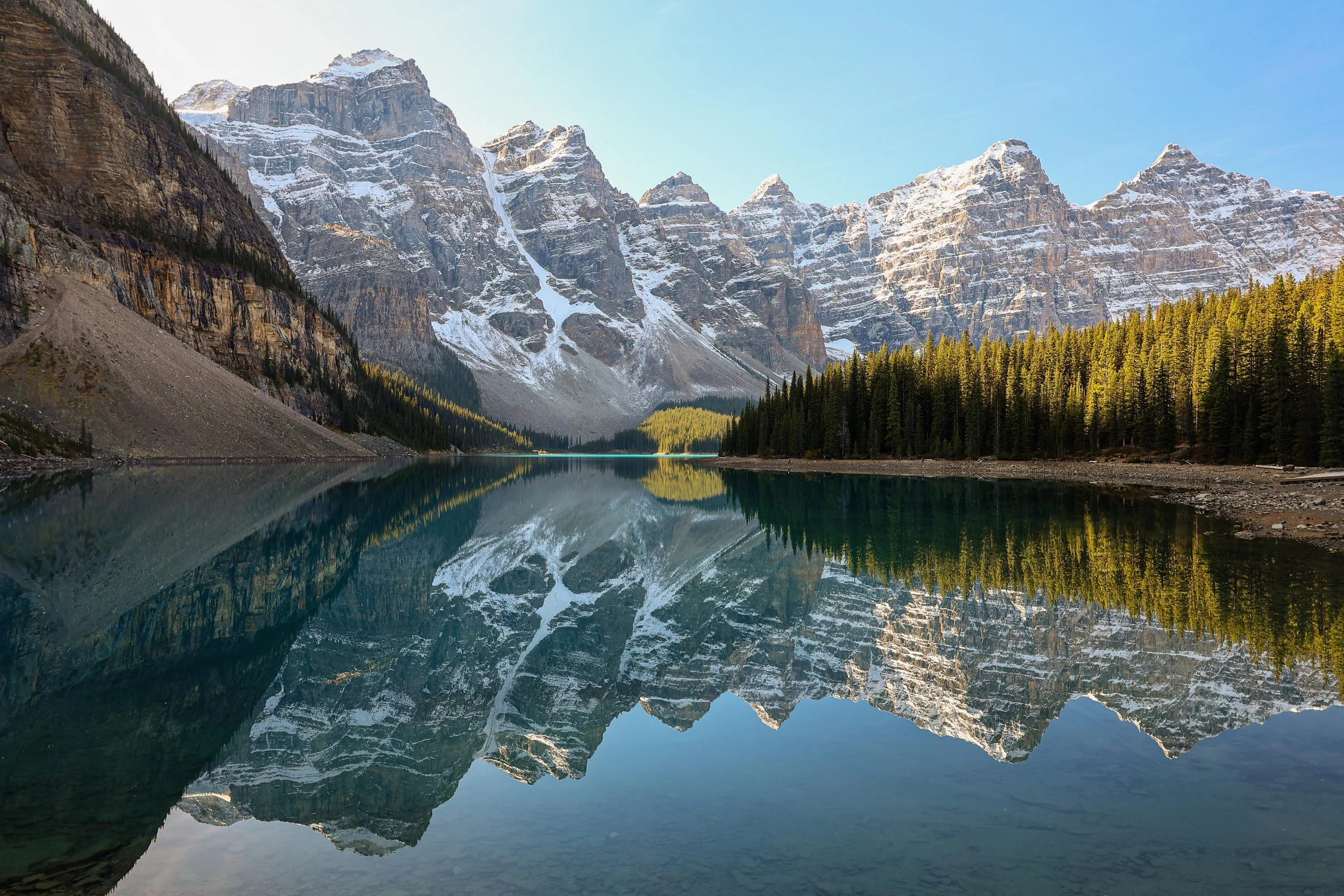Mountain range with snow-capped peaks reflected in a calm lake, surrounded by pine trees under a clear blue sky.