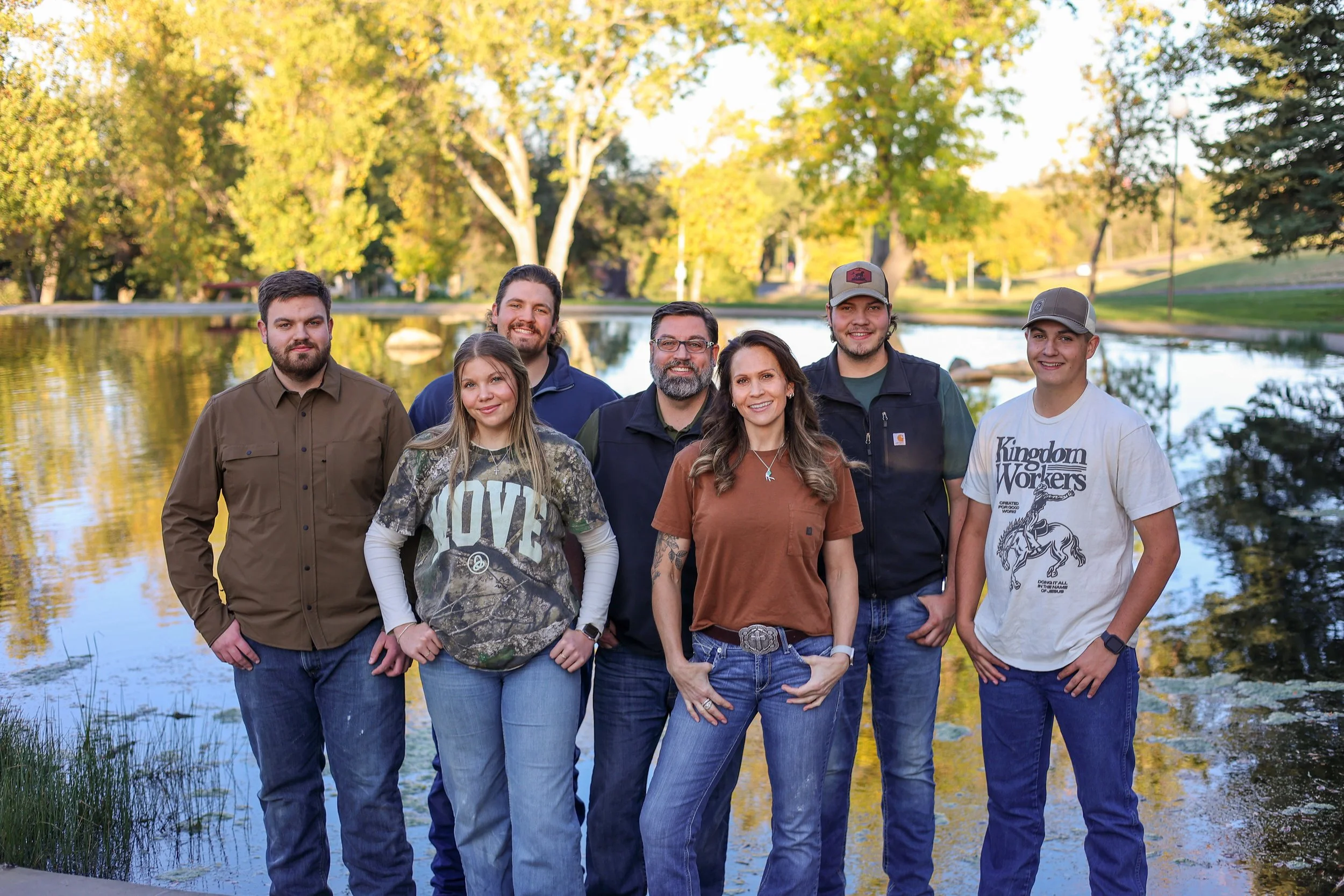 Group of seven people standing by a lake in a park during fall, smiling at the camera, with trees and reflections in the water.