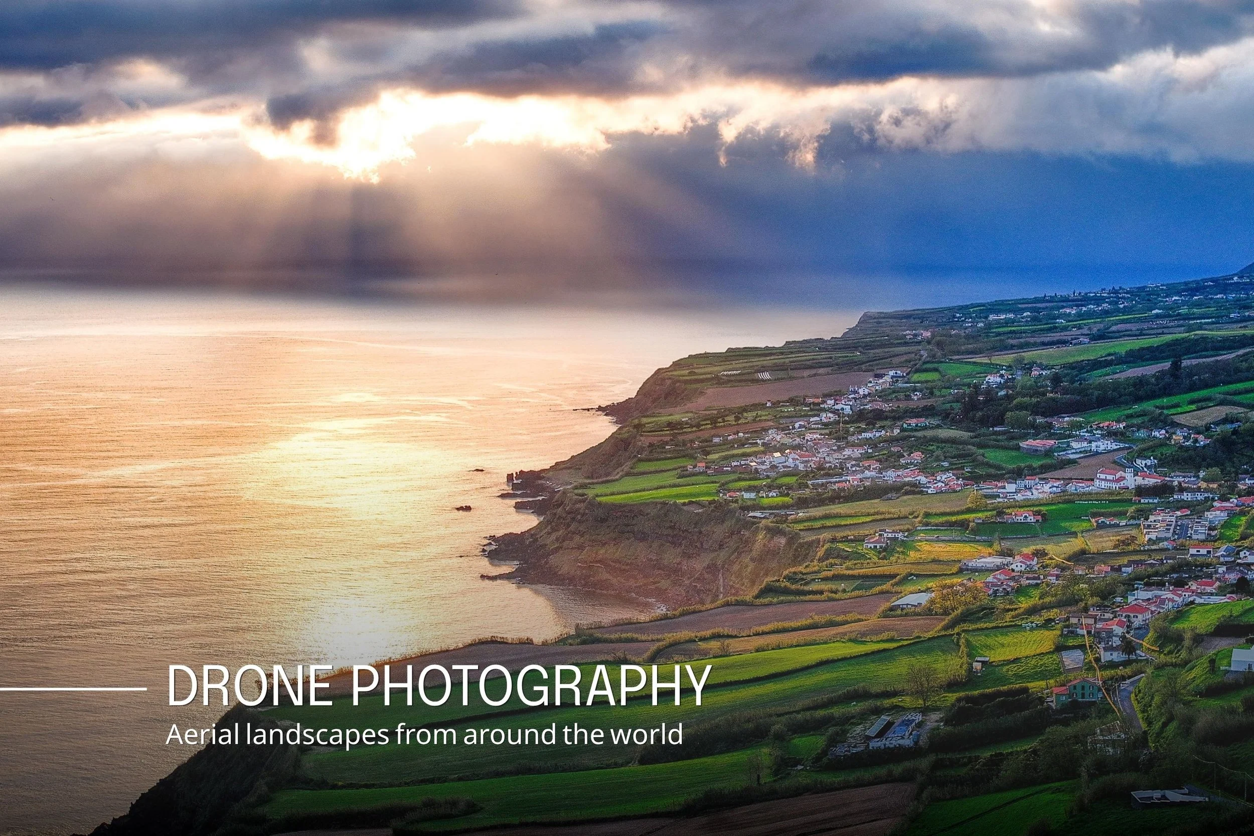 Aerial view of coastal landscape with cliffs, ocean, cloudy sky, and small town with houses and farmland.
