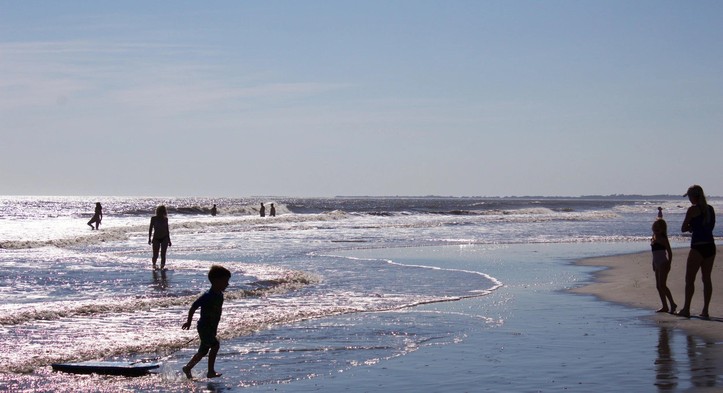 People enjoying the ocean at the beach on a sunny day, with children playing and surfboards in the water.