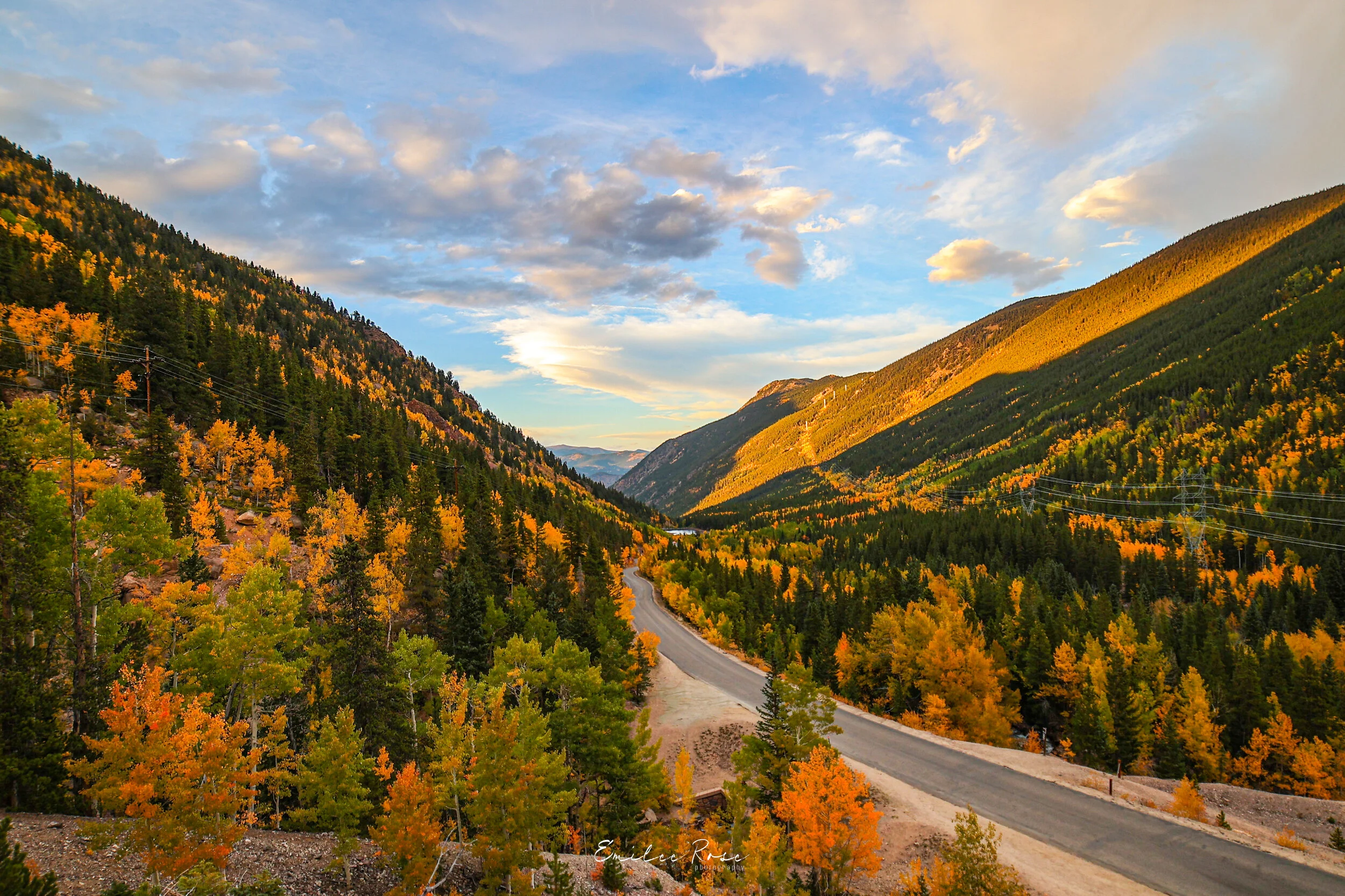 Scenic view of a mountain valley with trees in vibrant fall colors and a winding road, under a partly cloudy sky during sunset.