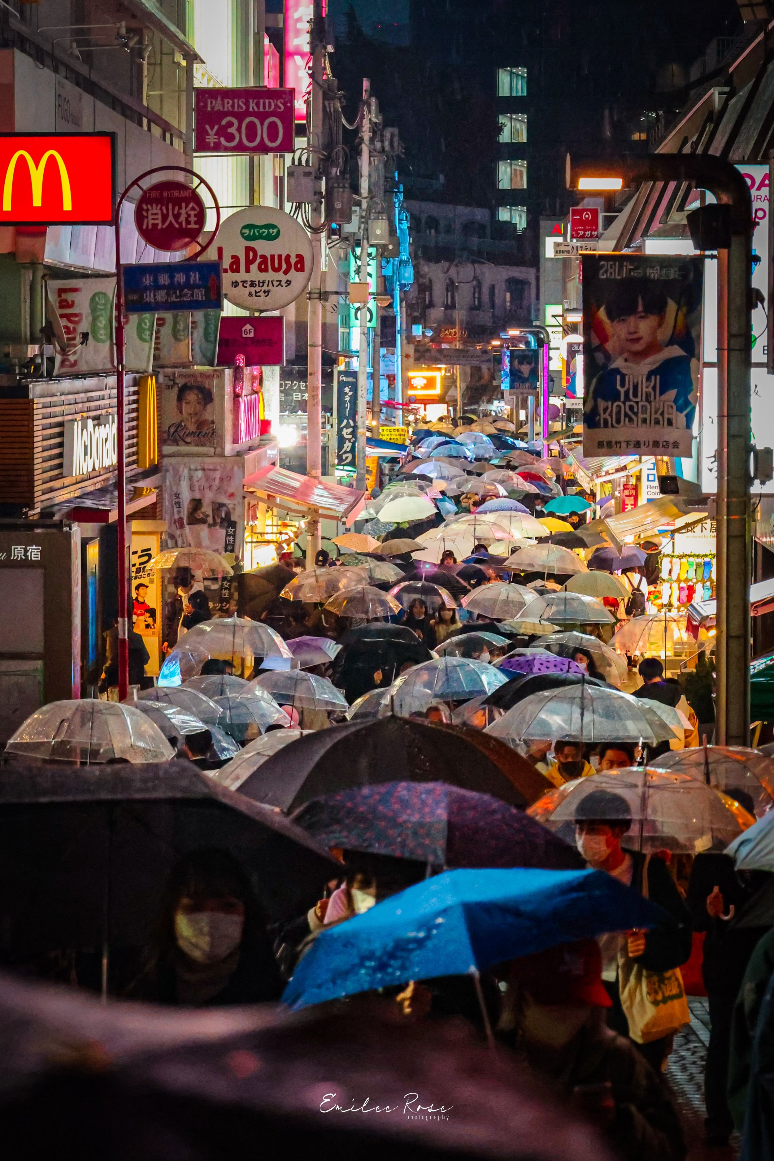 A crowded city street at night with many people holding umbrellas. Bright neon signs and advertisements are visible, creating a vibrant atmosphere, possibly in Japan.