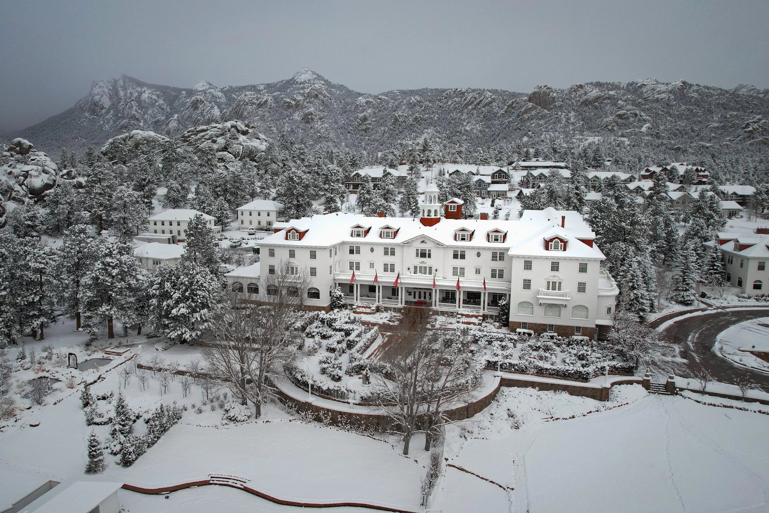 Snow-covered mountains in the background with a residential area of houses and a large white building with red rooftops in the foreground. Snow blankets the trees, rooftops, and ground.