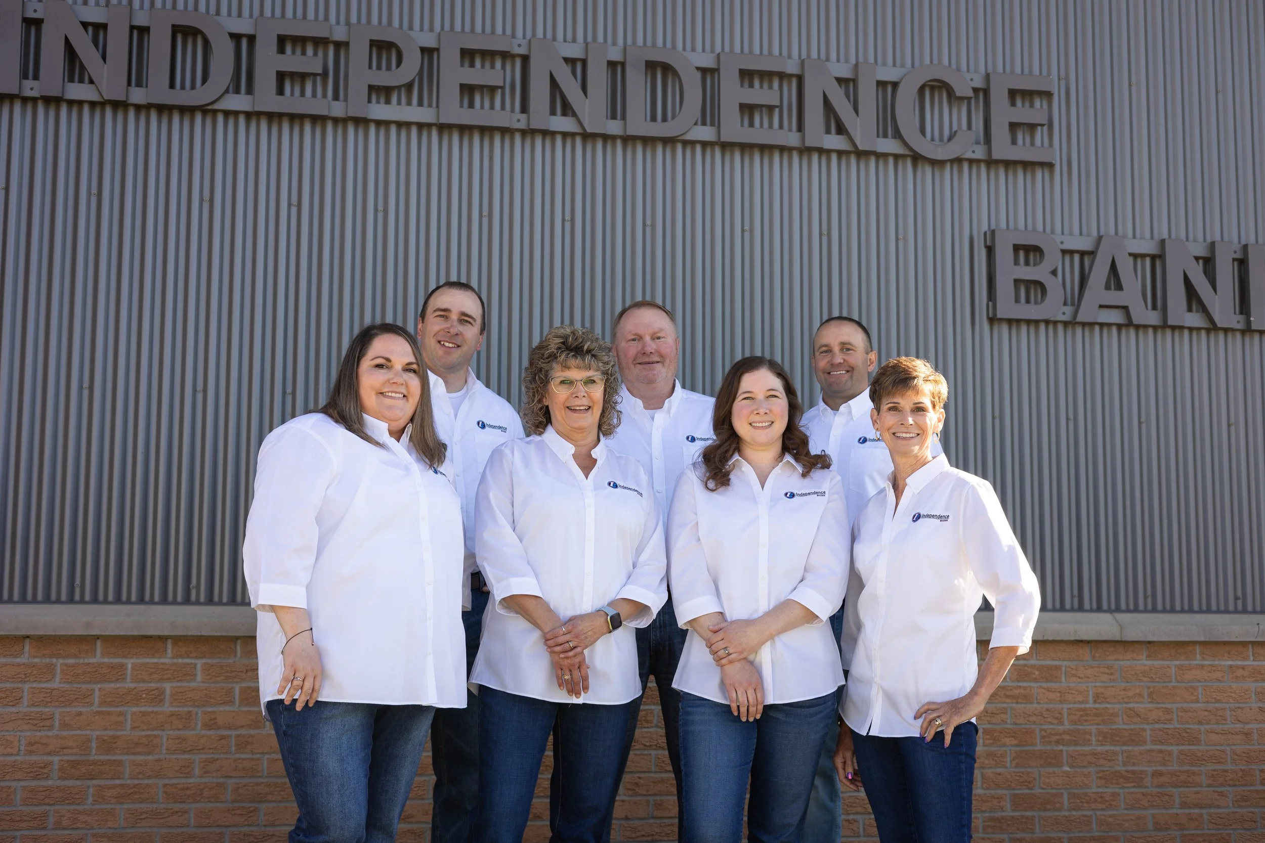 Group of seven people standing in front of a building with the words 'Independence Bank' on it, wearing white shirts and jeans, smiling at the camera.