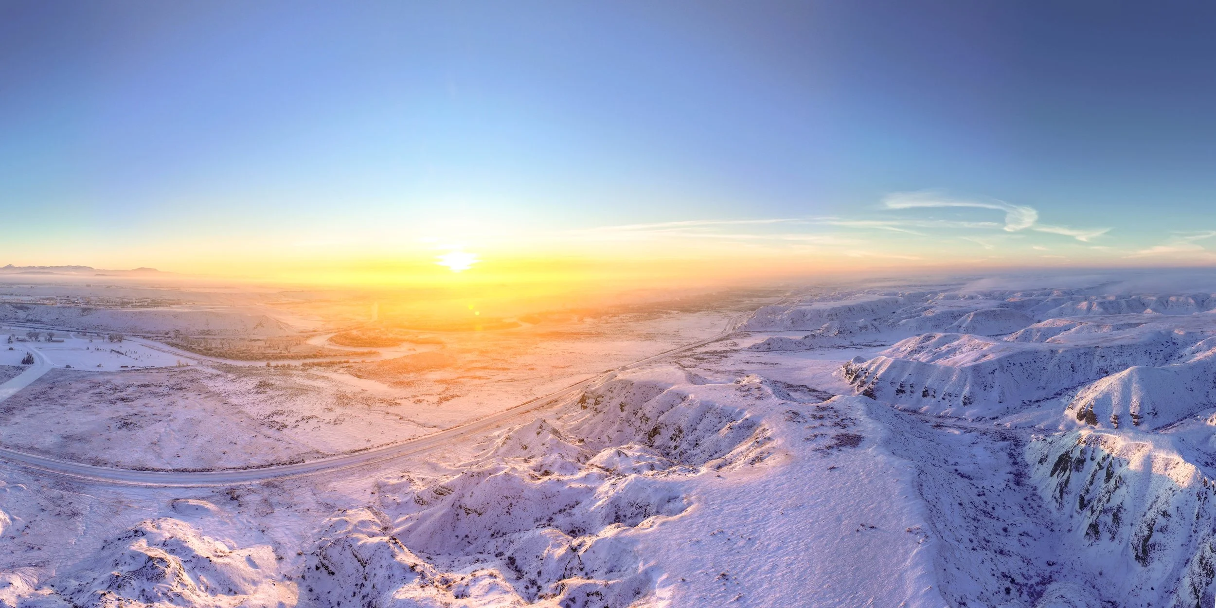 A panoramic view of snow-covered mountains and a vast flat valley during sunrise, with the sun near the horizon casting a warm glow over the landscape.