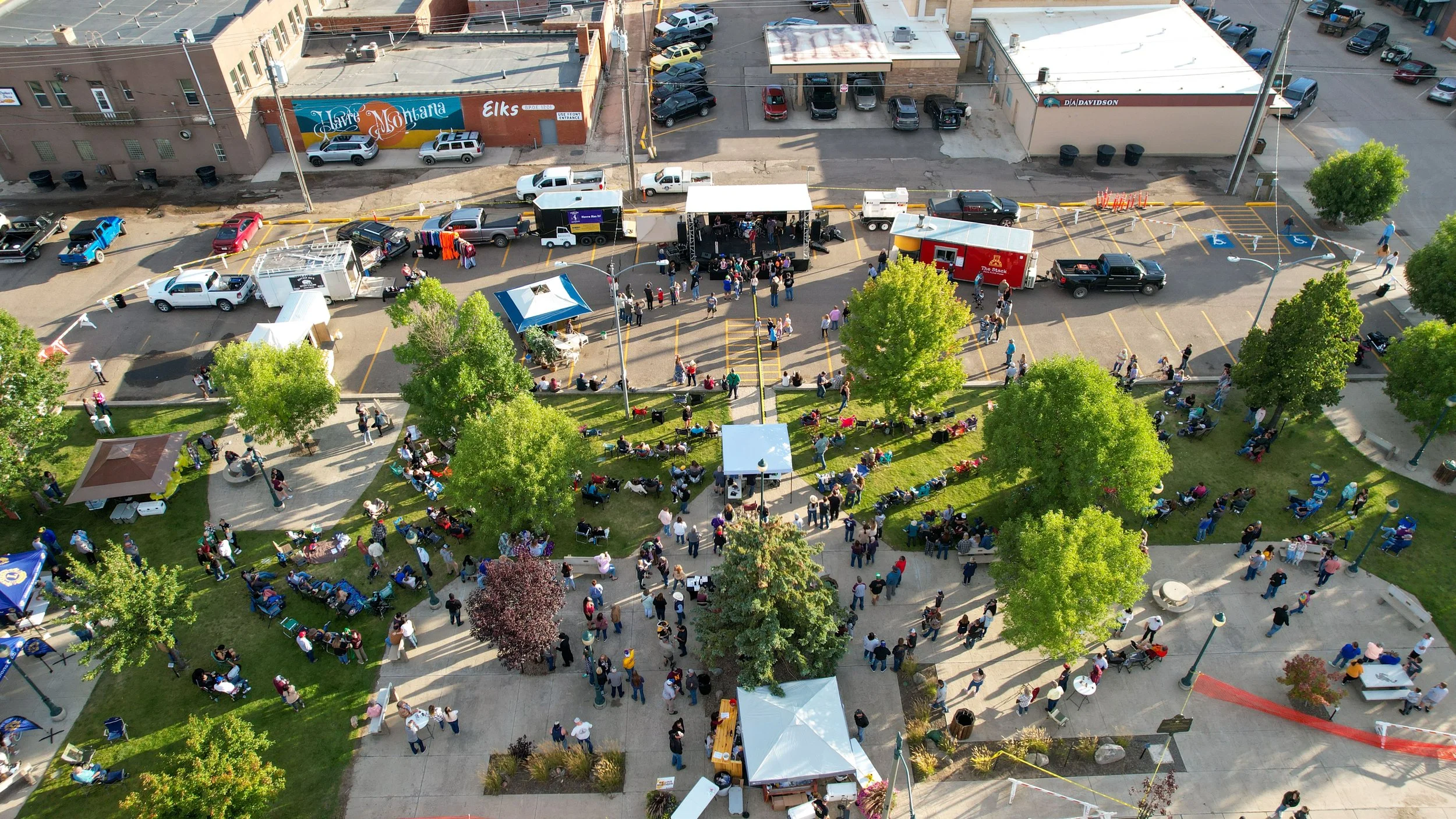 An aerial view of a community event in a park, featuring a stage with musicians performing, food trucks, tents, and many people sitting on benches, walking, and socializing among green trees and paved pathways.
