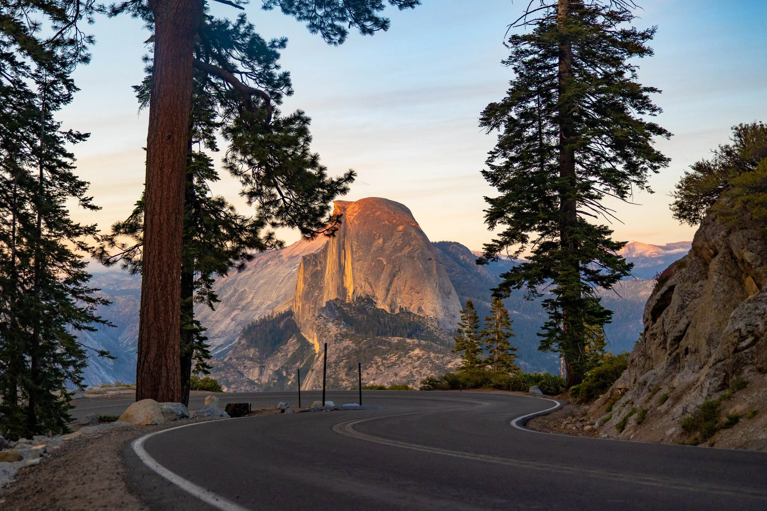 Scenic mountain landscape with tall trees, a winding road, and a prominent granite dome in the background, illuminated by sunset light.