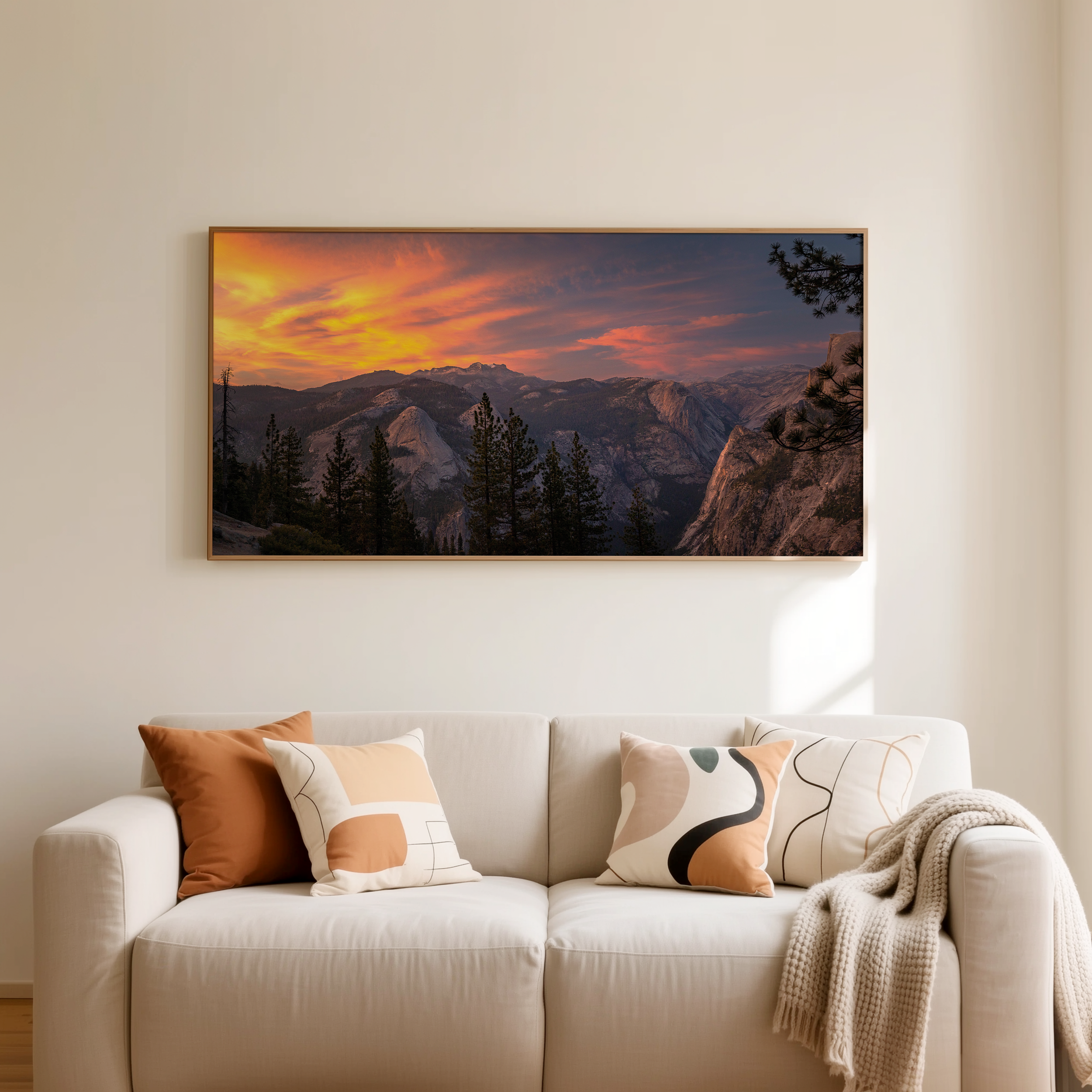 A sofa with multiple decorative pillows of various colors and patterns in a living room with a large framed landscape photograph of mountains and a sunset on the wall behind.