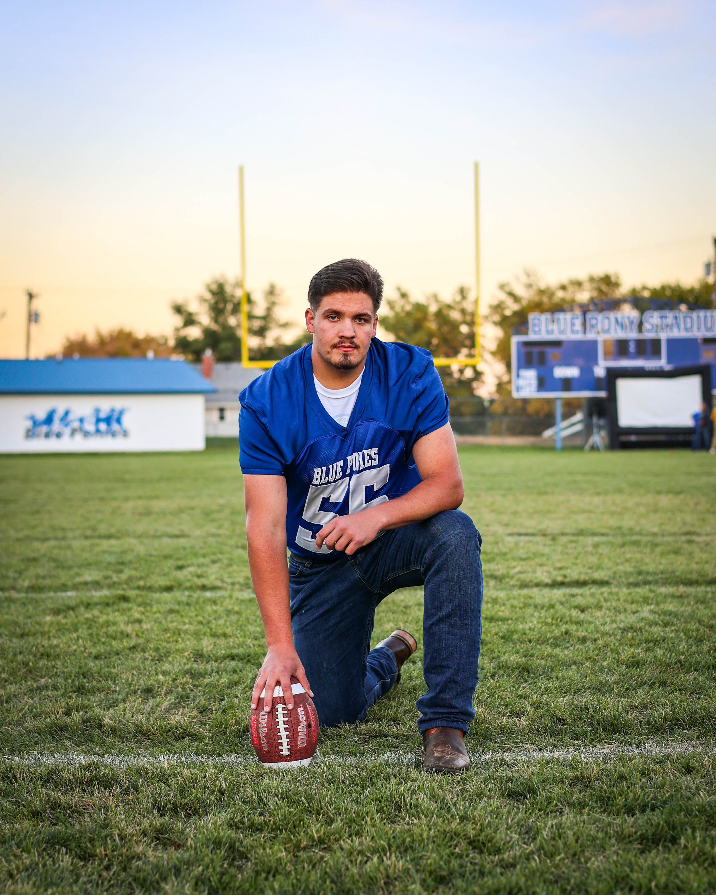 A young man kneeling on a football field holding a football with a stadium in the background.