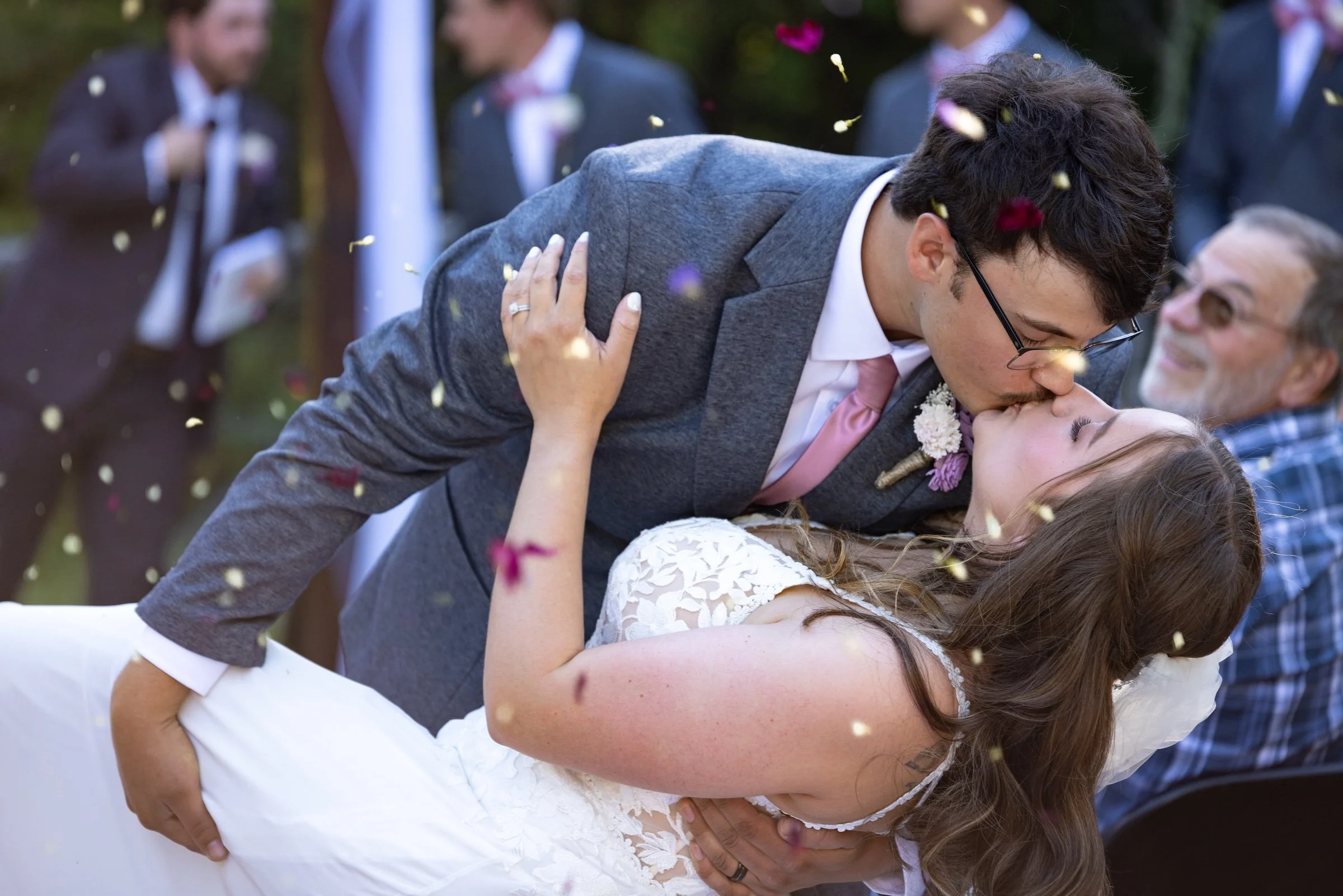 A newlywed couple sharing a kiss during their outdoor wedding celebration, with guests in the background and flower petals in the air.