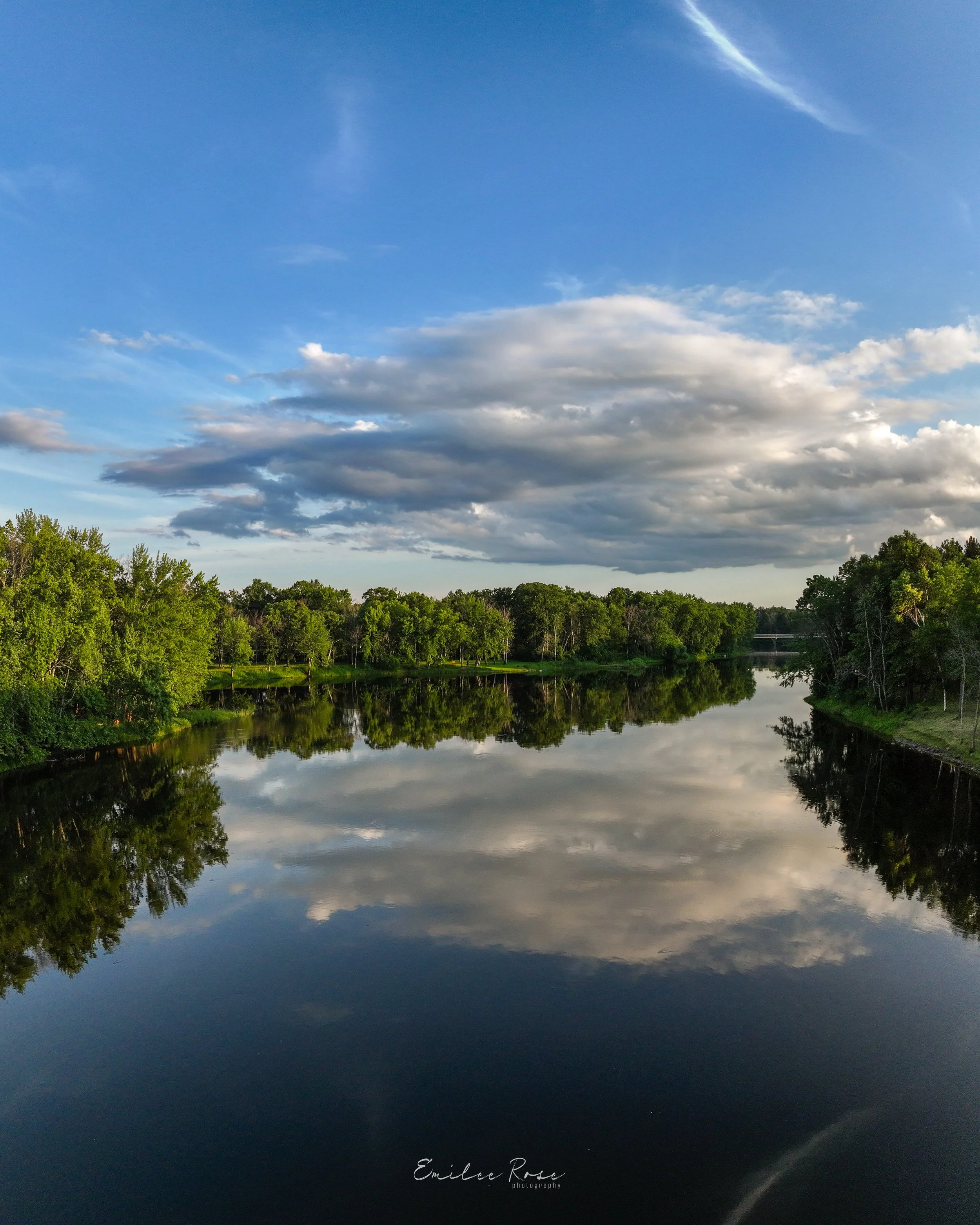 Calm river reflecting blue sky with white clouds and surrounding green trees.