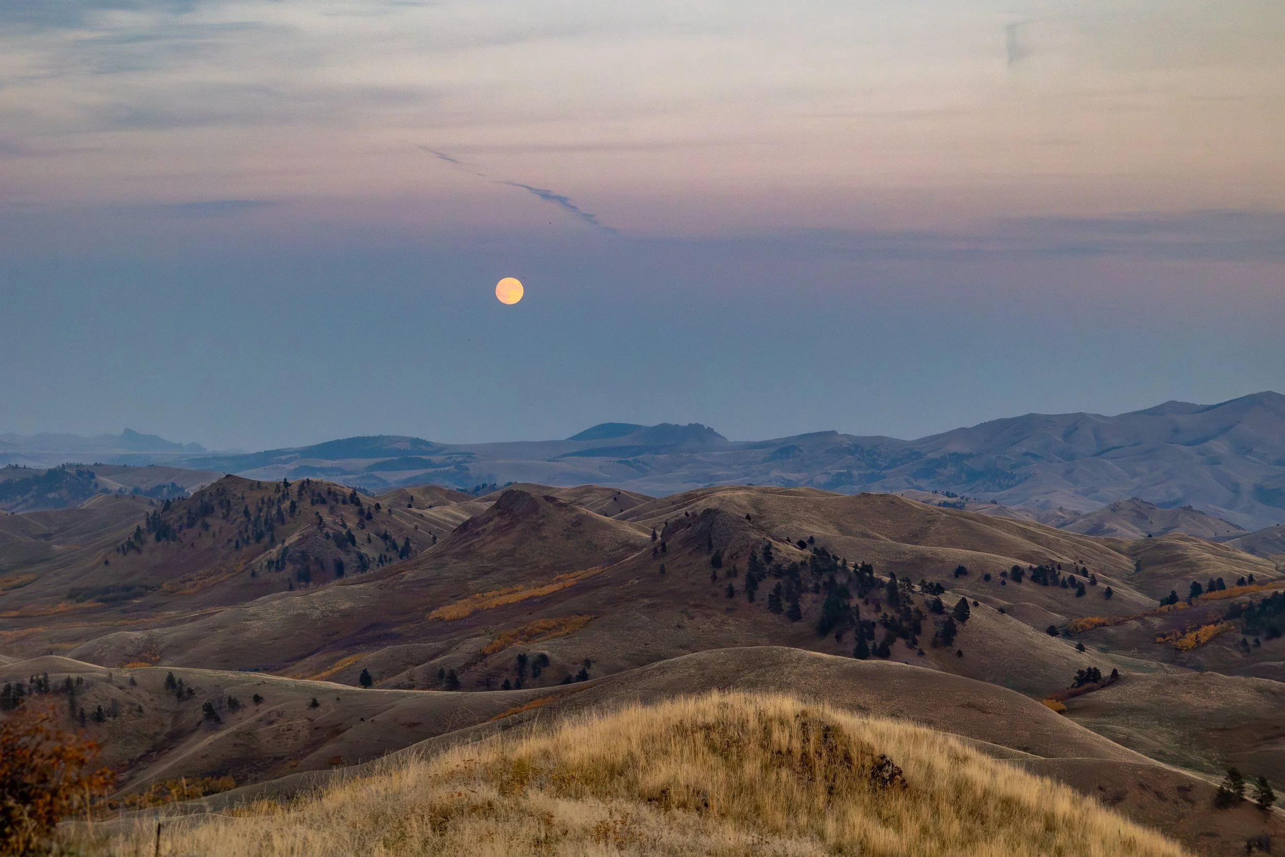 Landscape of rolling hills under a cloudy sky with the moon visible near the horizon.