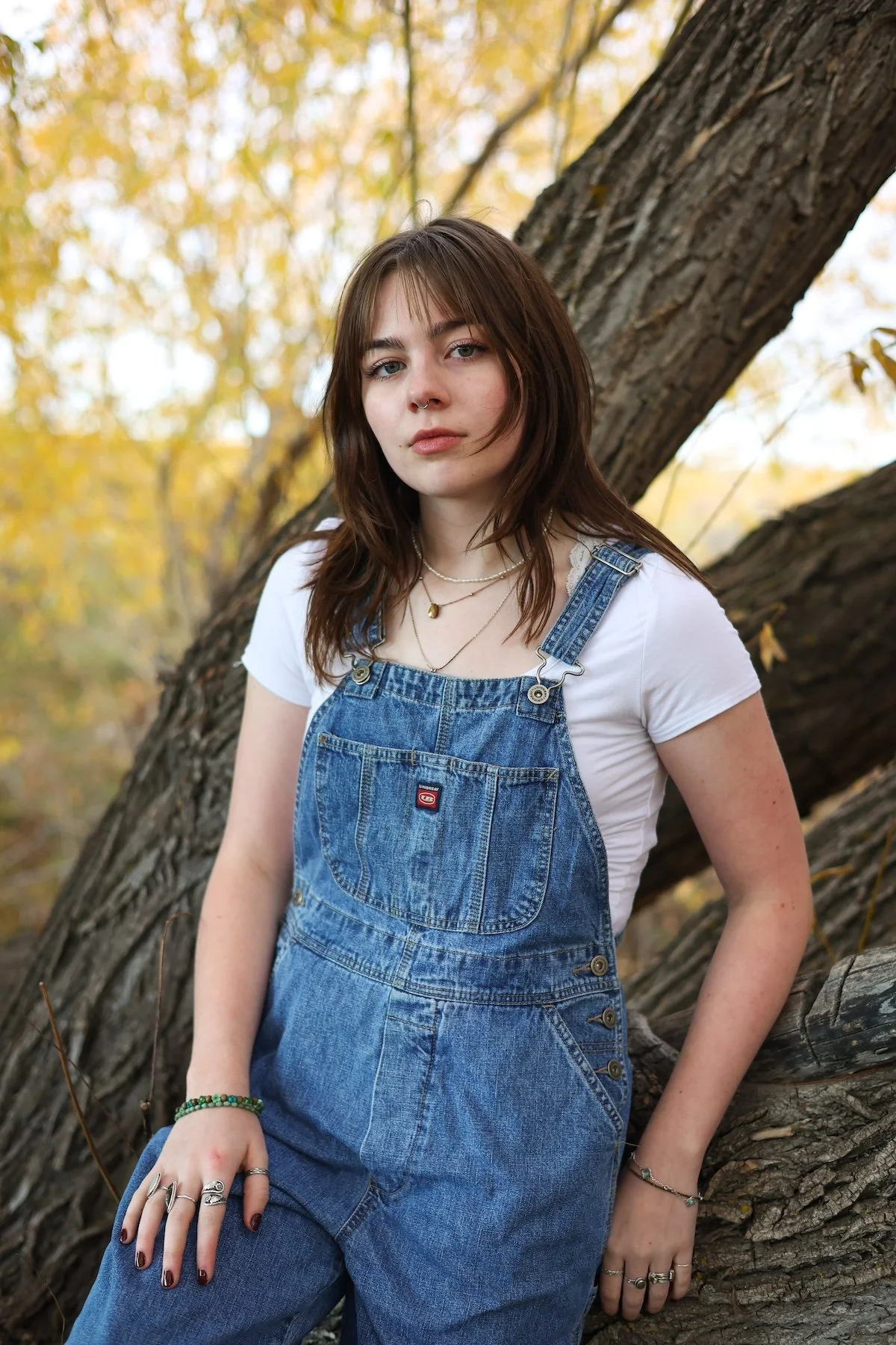 A young woman with brown hair and a septum piercing standing outdoors near a large tree with yellow leaves during autumn.