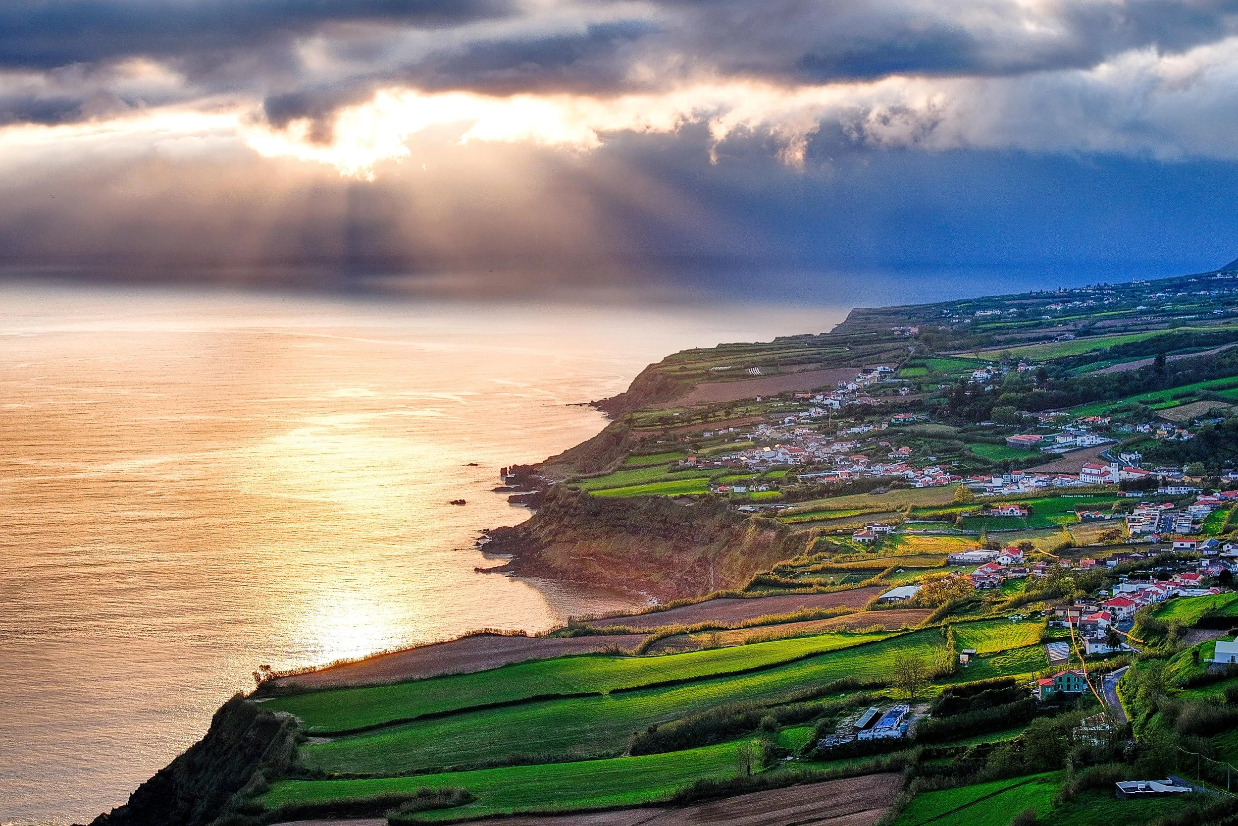 Sunset over a coastal village with green fields, houses, and a steep cliff with dark clouds and sun rays shining through.