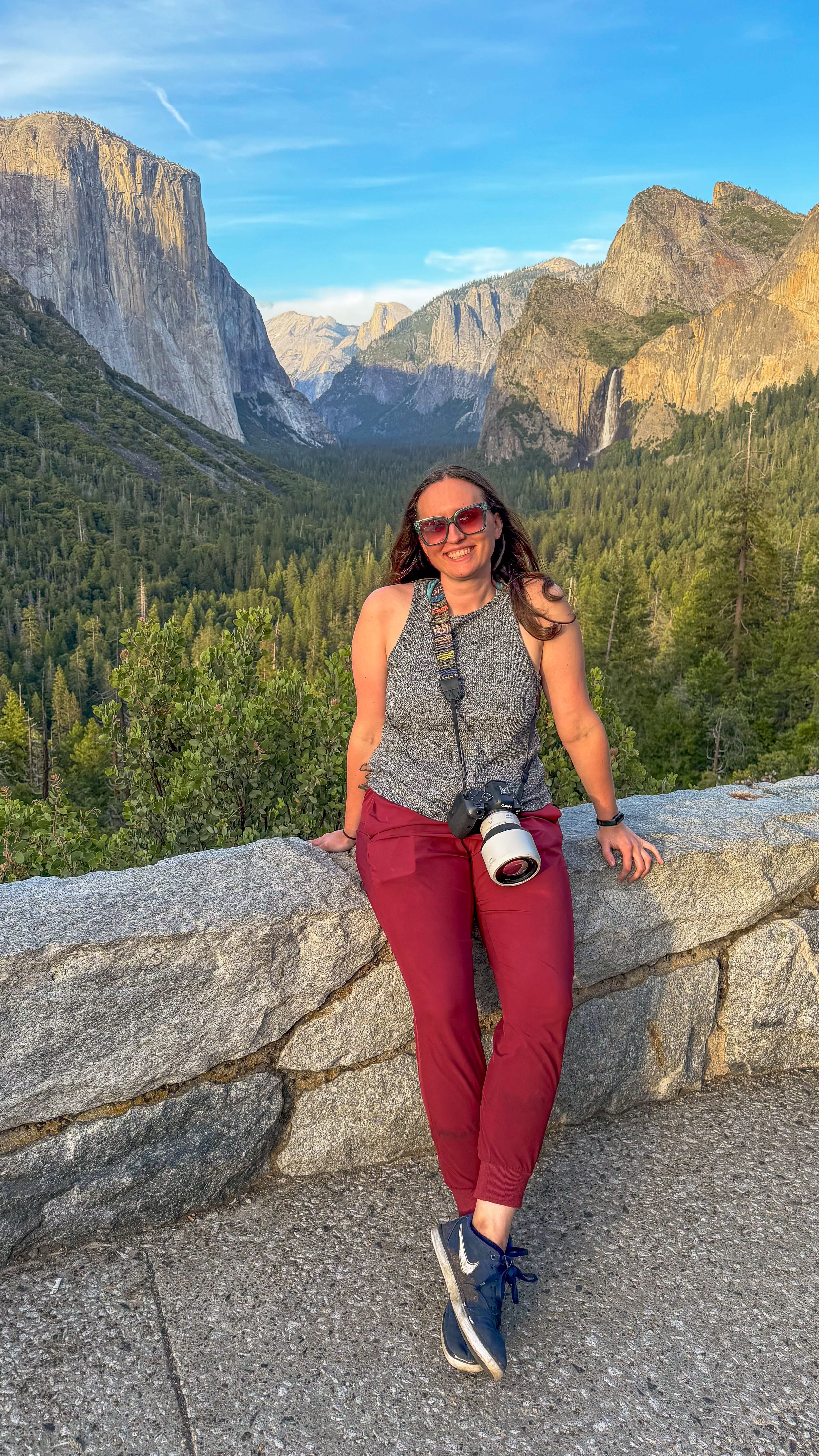 A woman smiling, wearing sunglasses, a sleeveless gray top, red pants, and black Nike sneakers, is sitting on a stone ledge in a mountainous forest area with cliffs and waterfalls in the background.