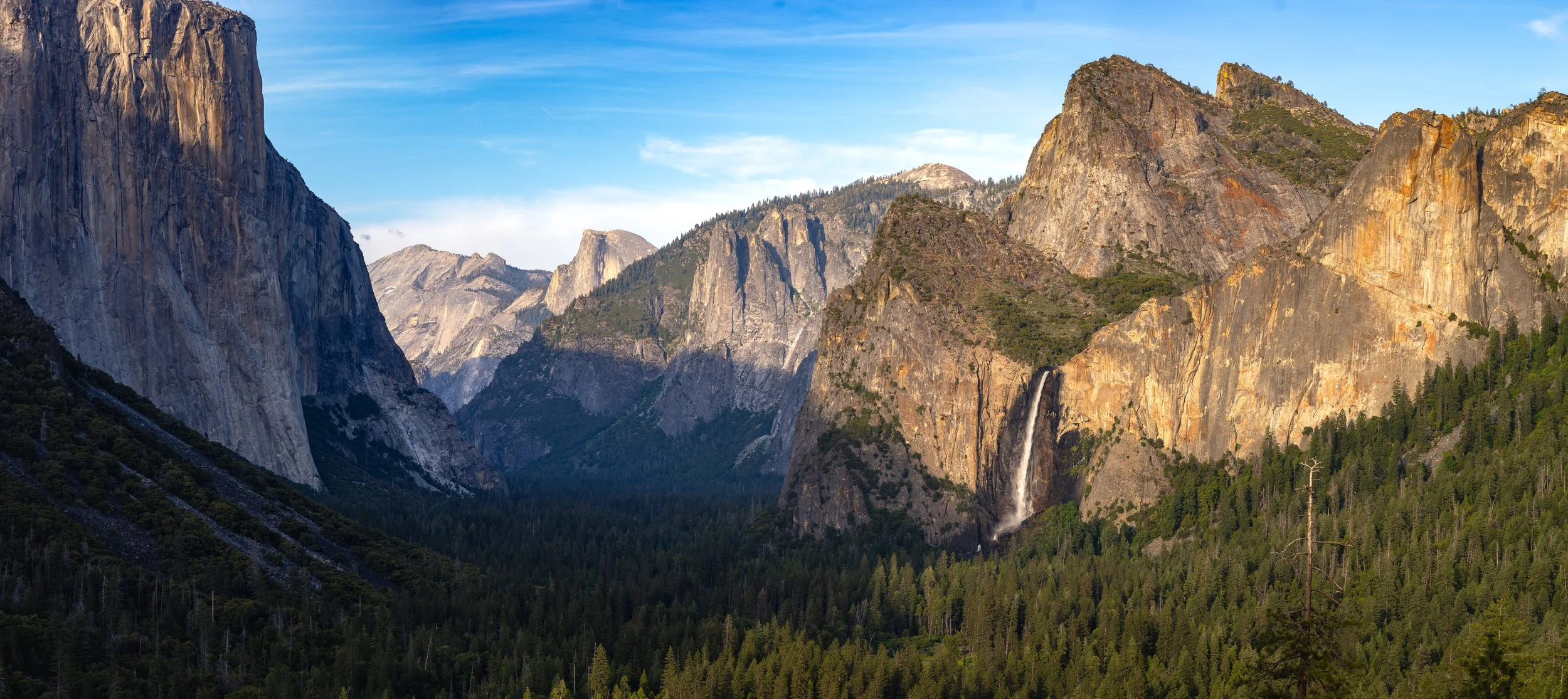 Scenic view of Yosemite National Park featuring towering granite cliffs, a waterfall, and dense green forests under a blue sky.