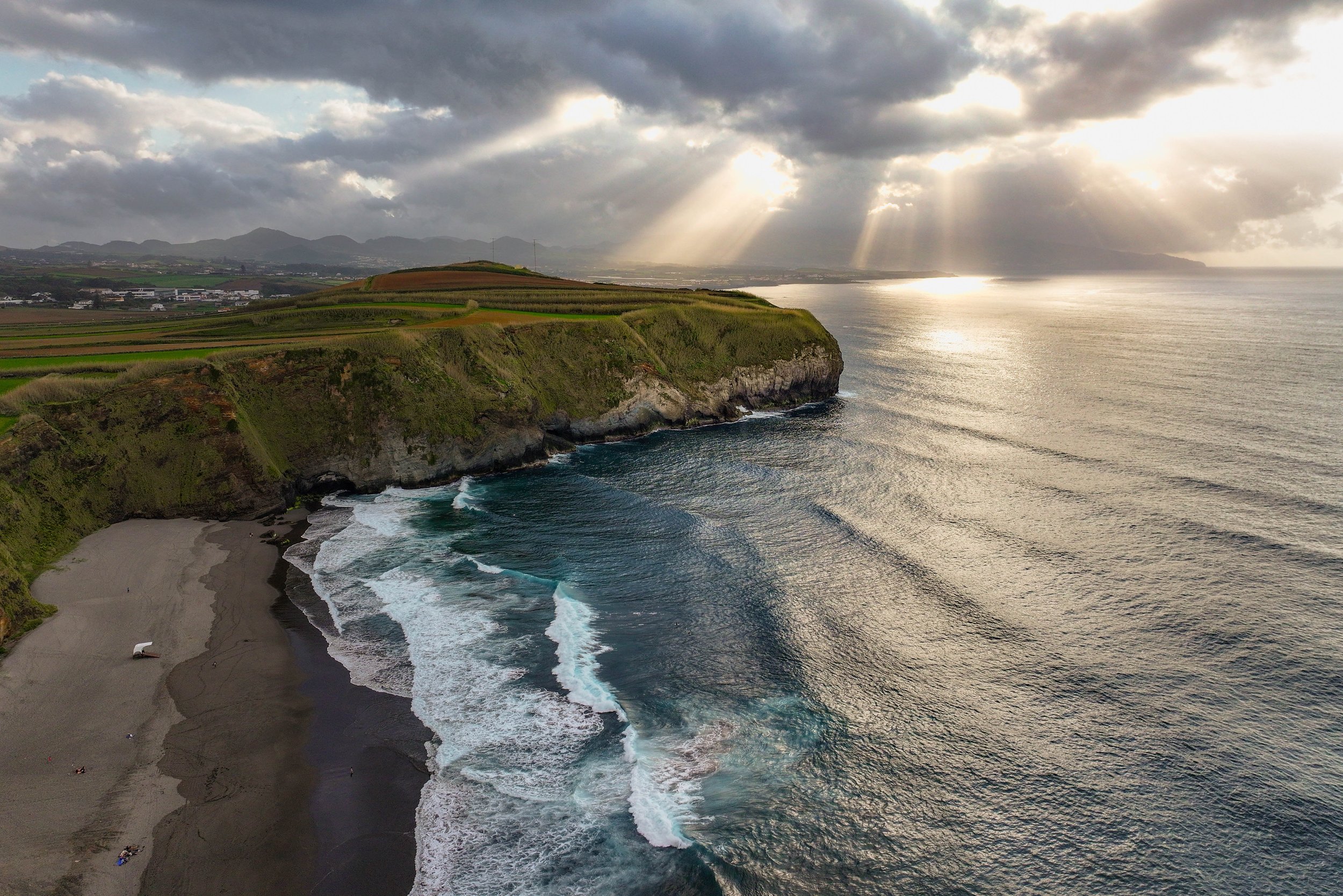 A coastline with rocky cliffs and black sand beach, waves crashing near the shore, farmland and mountains in the background under a cloudy sky with rays of sunlight breaking through.