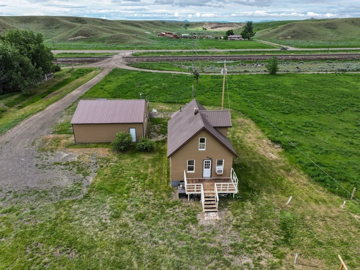 A small house with a porch and stairs is surrounded by green grassy fields and a separate garage or shed nearby. In the background, a highway, train tracks, and rolling hills under a cloudy sky are visible.