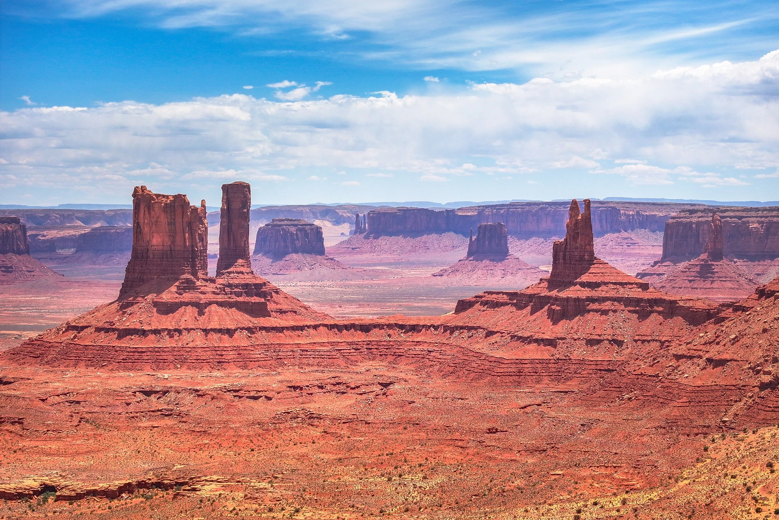 A landscape of the desert with red rock formations and buttes under a partly cloudy blue sky.