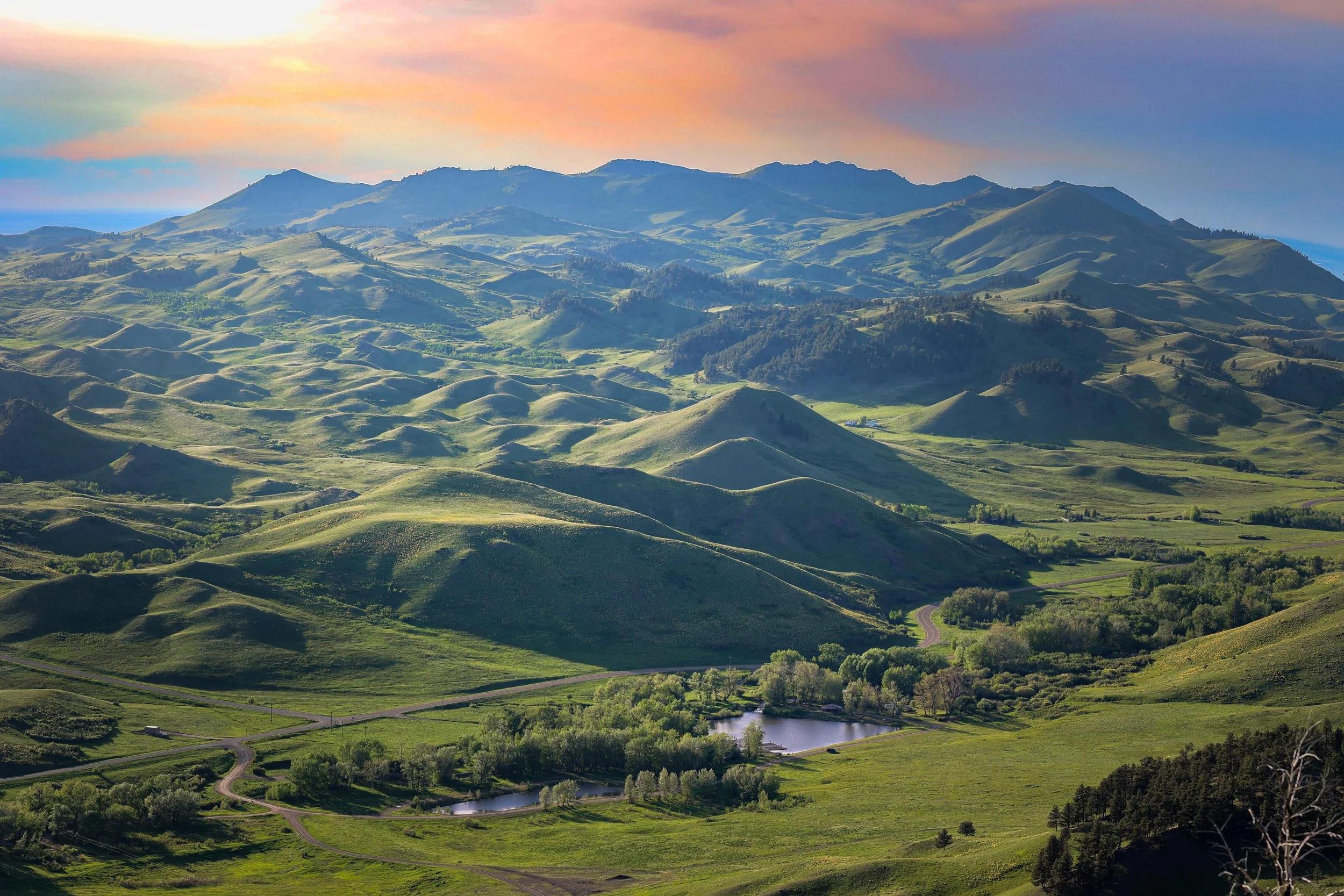 Scenic view of rolling green hills and mountains during sunset with a sky painted in soft pastel colors, and a winding road visible among the landscape.