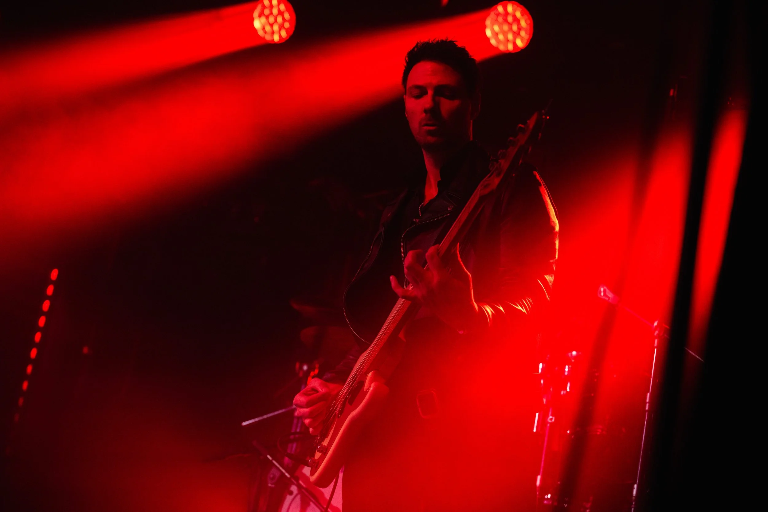 Man playing an electric guitar on stage, illuminated by red stage lighting and background lights.
