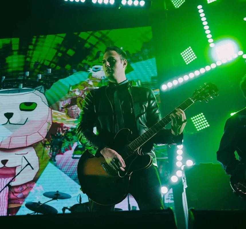 Musician playing guitar on stage with green lighting and digital artwork in background.