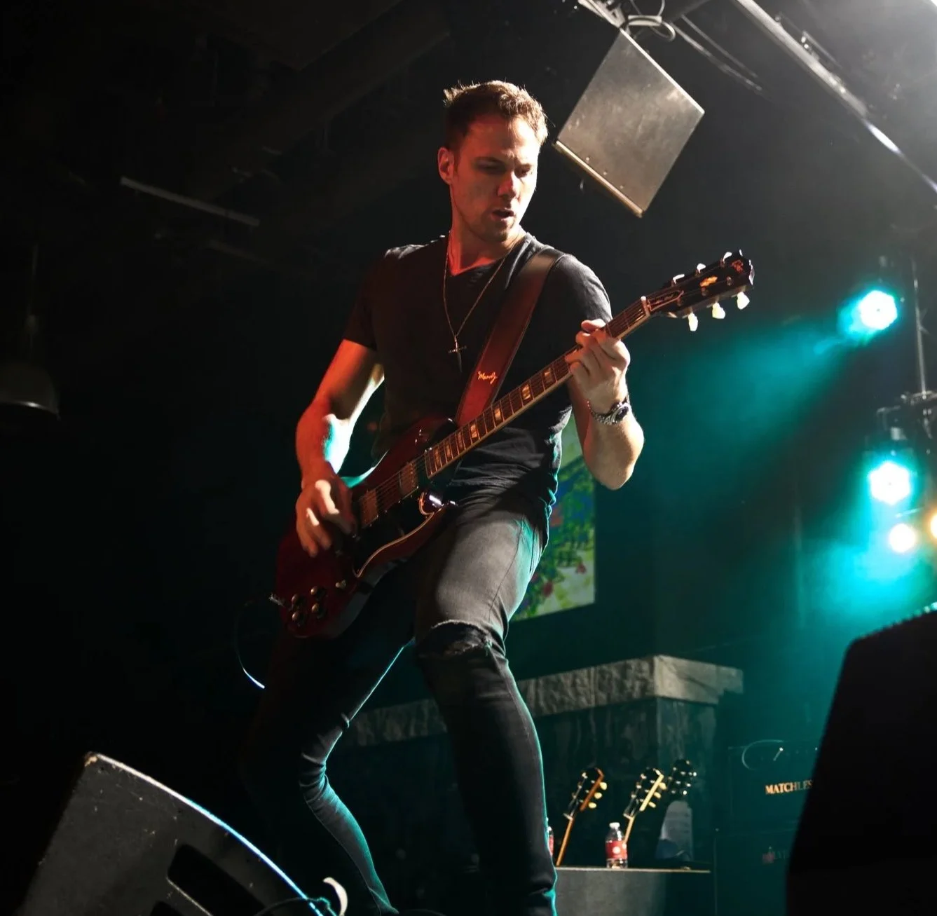 A male guitarist playing an electric guitar on stage with colorful stage lights in the background.