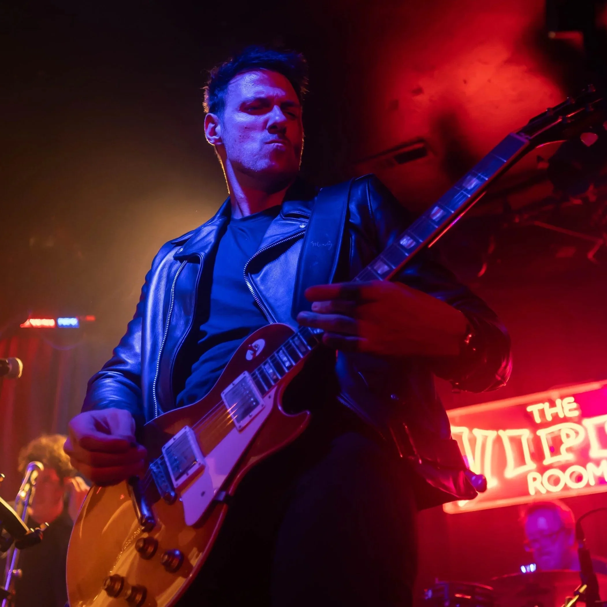 Music performer playing electric guitar on stage with red and purple lighting.