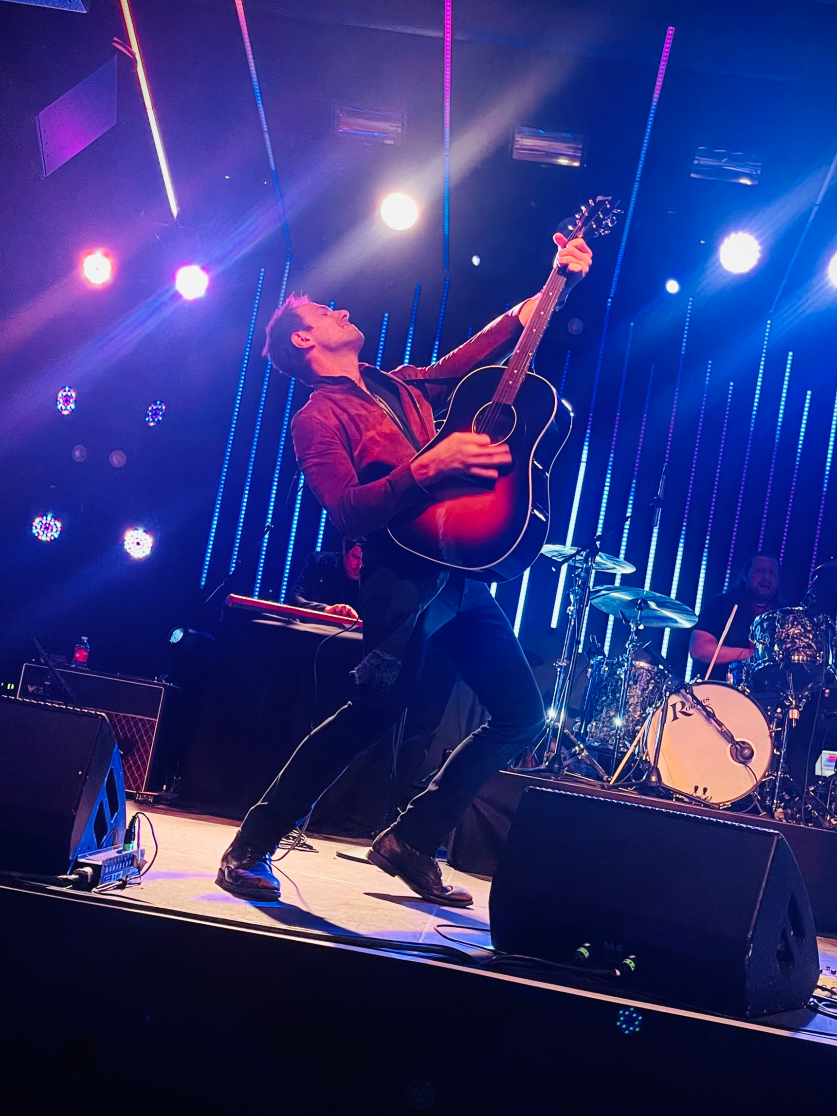 Musician playing guitar on stage with colorful vertical lights and stage spotlights, drummer visible in the background.