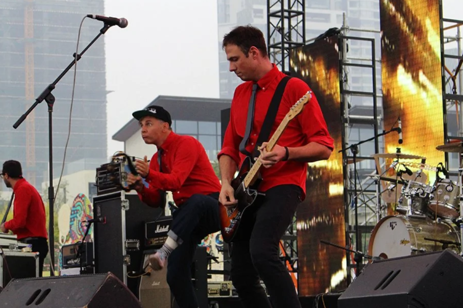 A band performing on an outdoor stage with city buildings in the background. The musicians are wearing red shirts; one is playing guitar, another is adjusting equipment, and a third is playing drums.