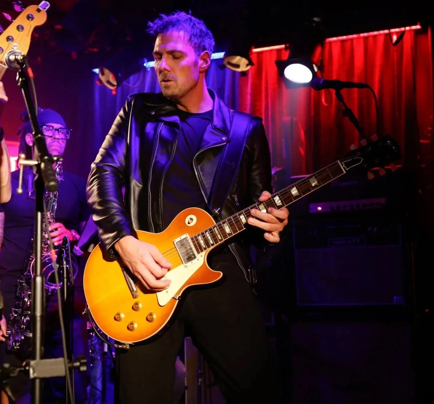 A male musician wearing a black leather jacket playing an orange electric guitar on stage with red curtains in the background.