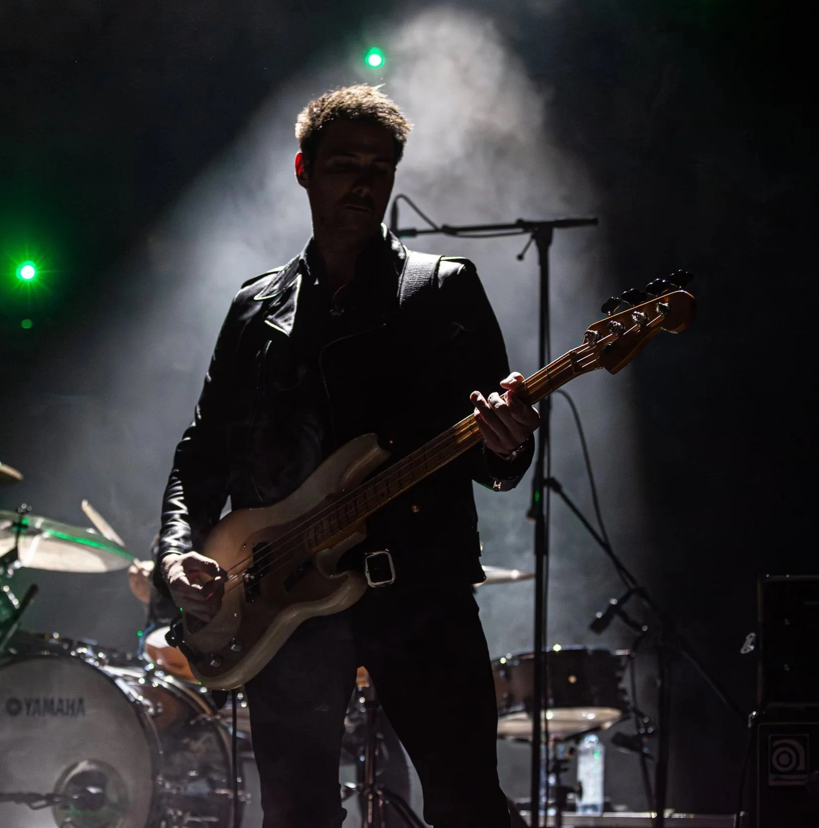 A musician in dark clothing playing an electric guitar on stage, with drum set and microphone in the background under stage lighting,