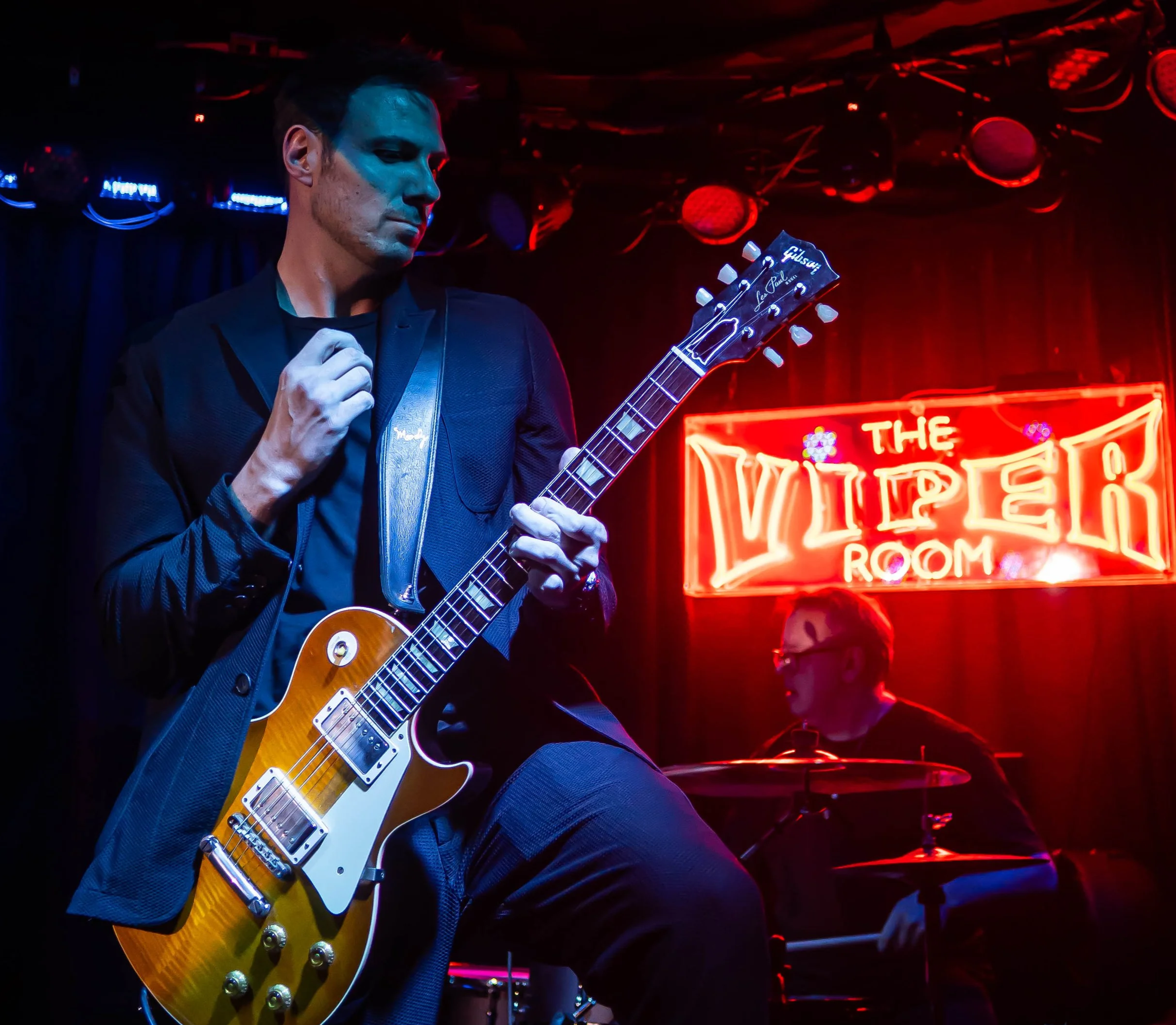 A man playing a yellow Gibson Les Paul electric guitar on stage in a dimly lit venue with red and blue lighting, and a neon sign reading 'The Viper Room' in the background.