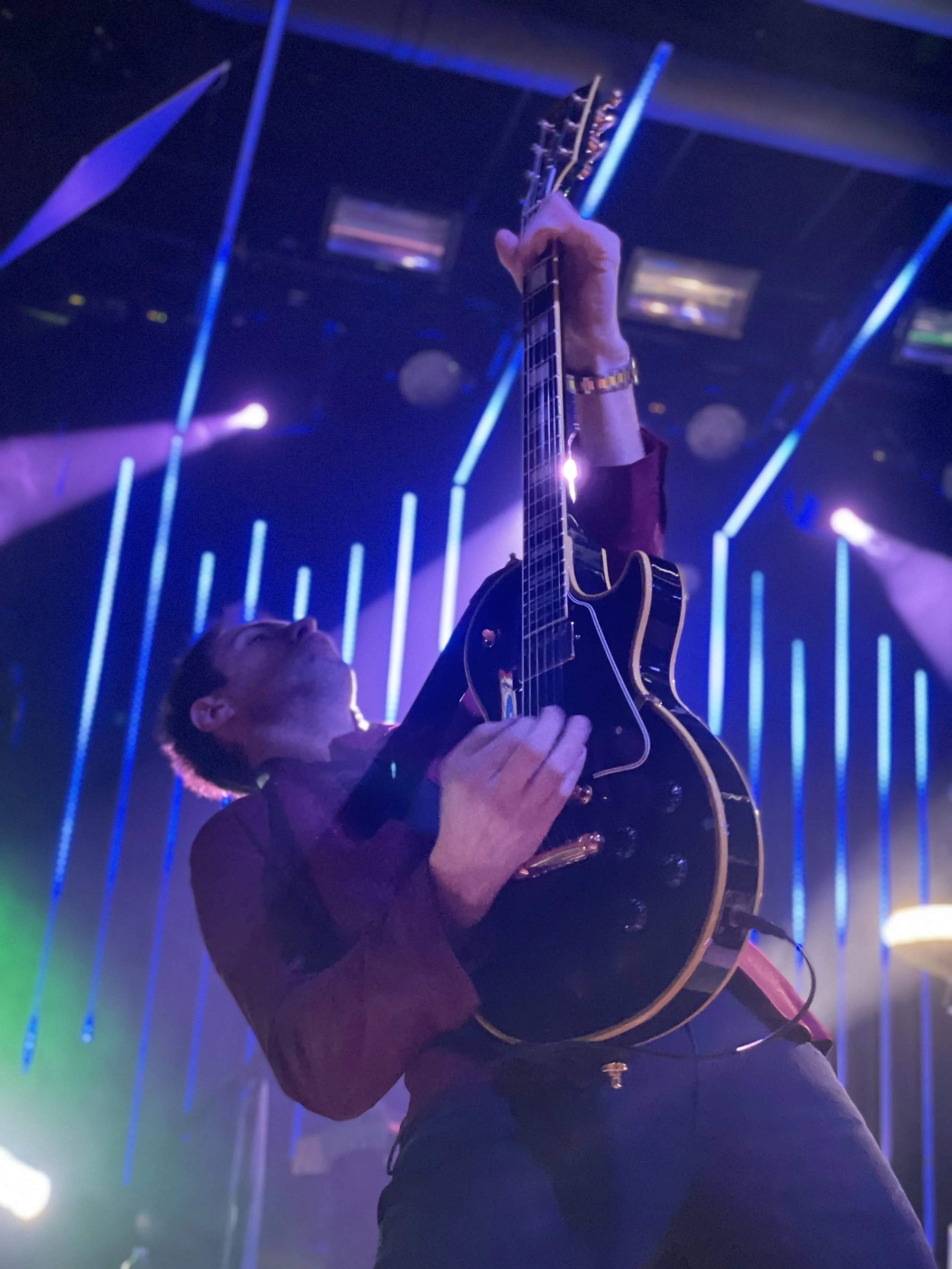 A man playing an electric guitar on stage with vibrant blue and purple lighting, and vertical LED light strips in the background. The man is wearing a dark shirt and a watch, and is captured in a close-up shot from a low angle.