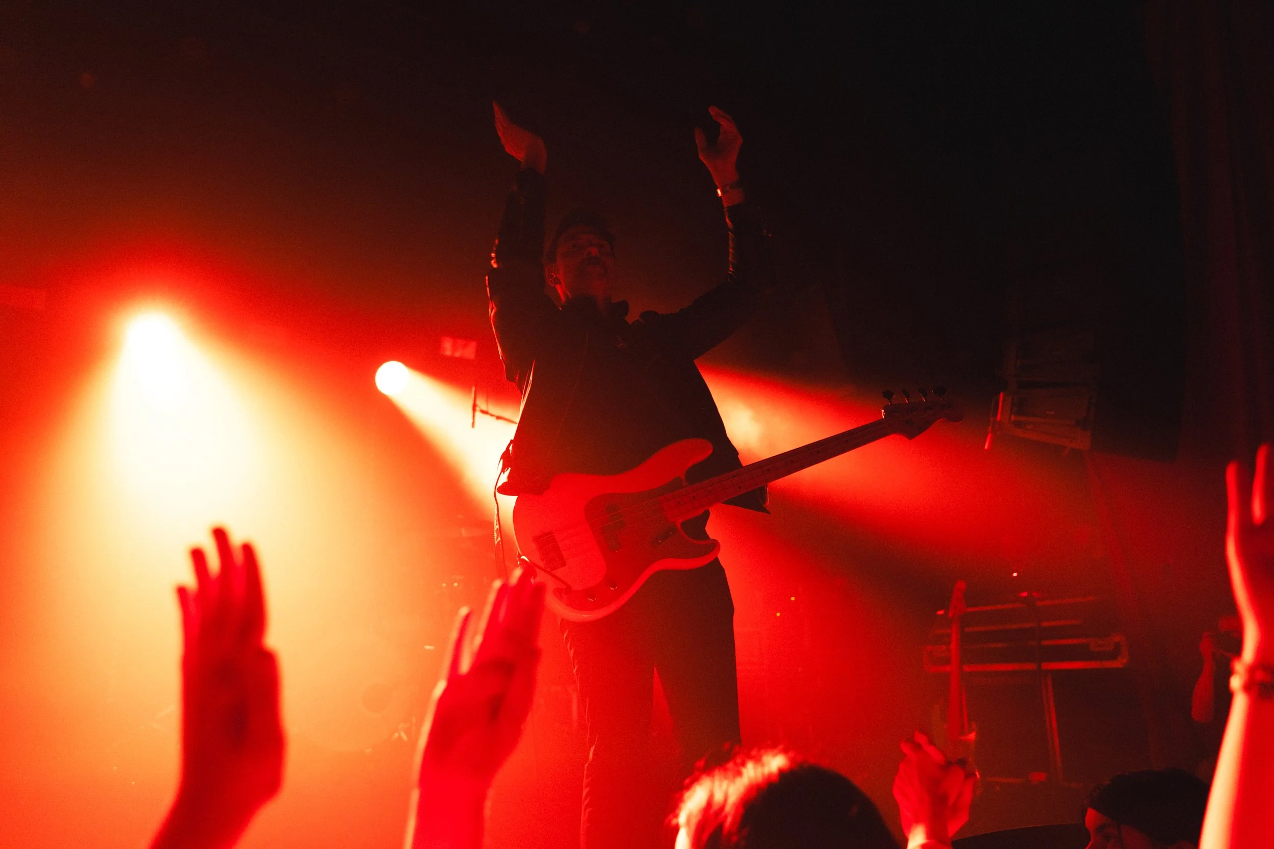 A musician playing electric guitar on stage illuminated by red lights, with hands raised in the audience.