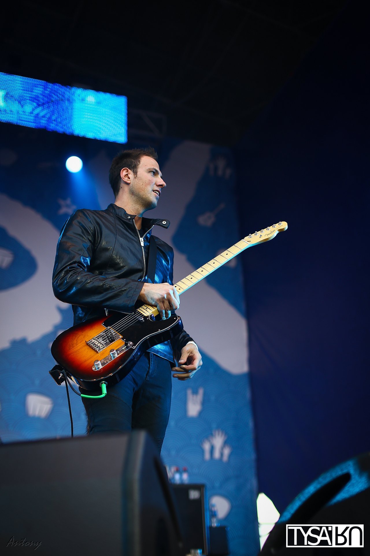 A male musician wearing a black leather jacket playing an electric guitar on stage. He appears to be concentrating, with a blue stage light overhead. The background has a stylized design and the TV5MONDE logo in the bottom right corner.