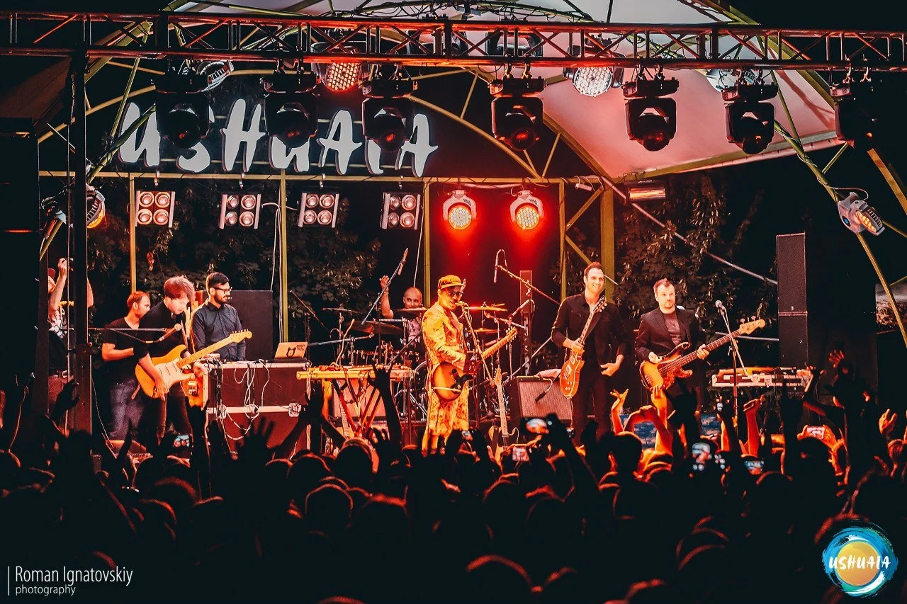 Live outdoor concert with a band performing on stage, audience members capturing the moment with their smartphones, stage lighting, and audience hands raised.