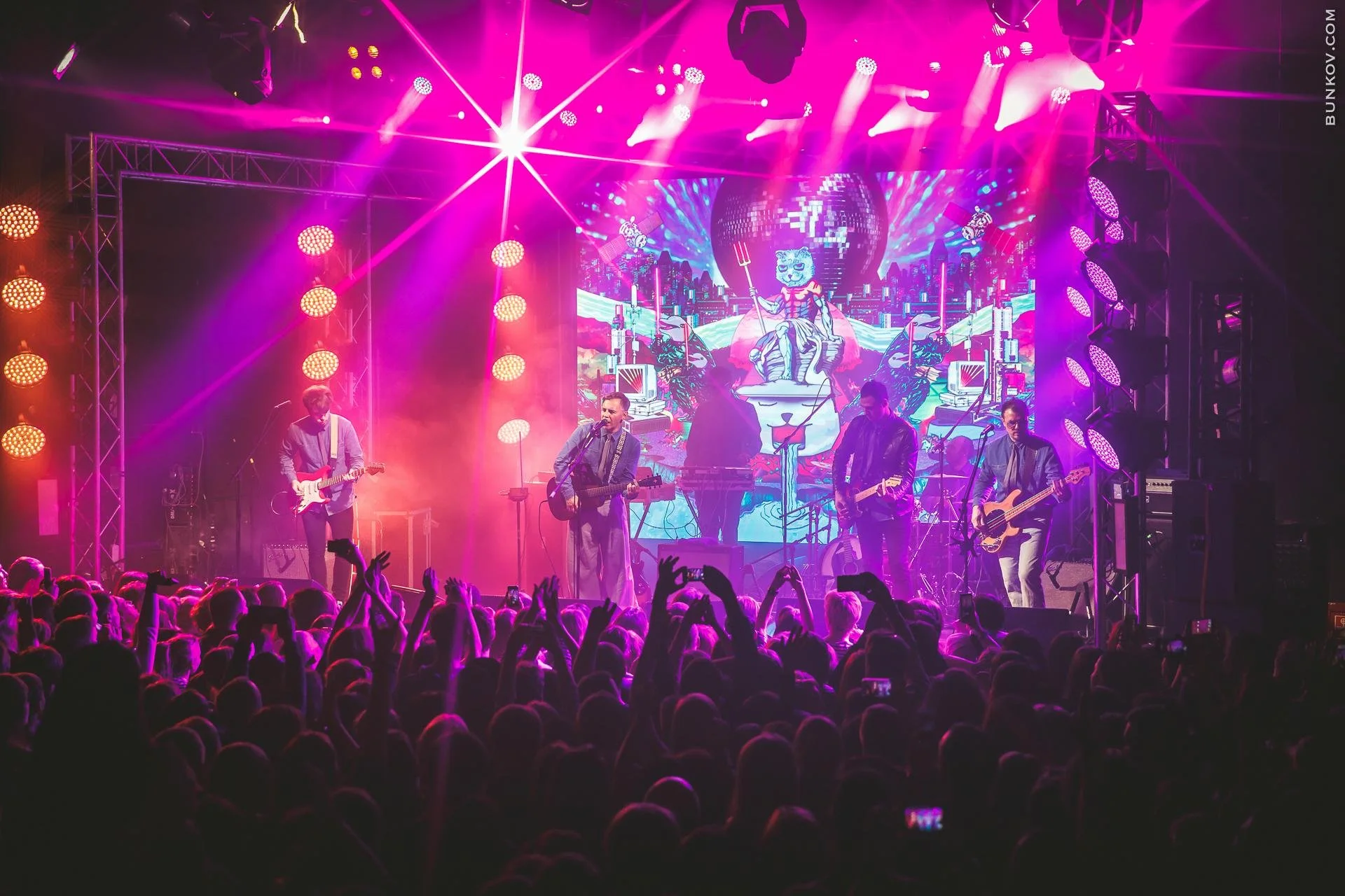 A concert stage with five musicians performing under colorful pink and purple lighting, with a large digital backdrop displaying a cartoonish tiger with a cityscape background. The audience is visible in the foreground with many hands raised.