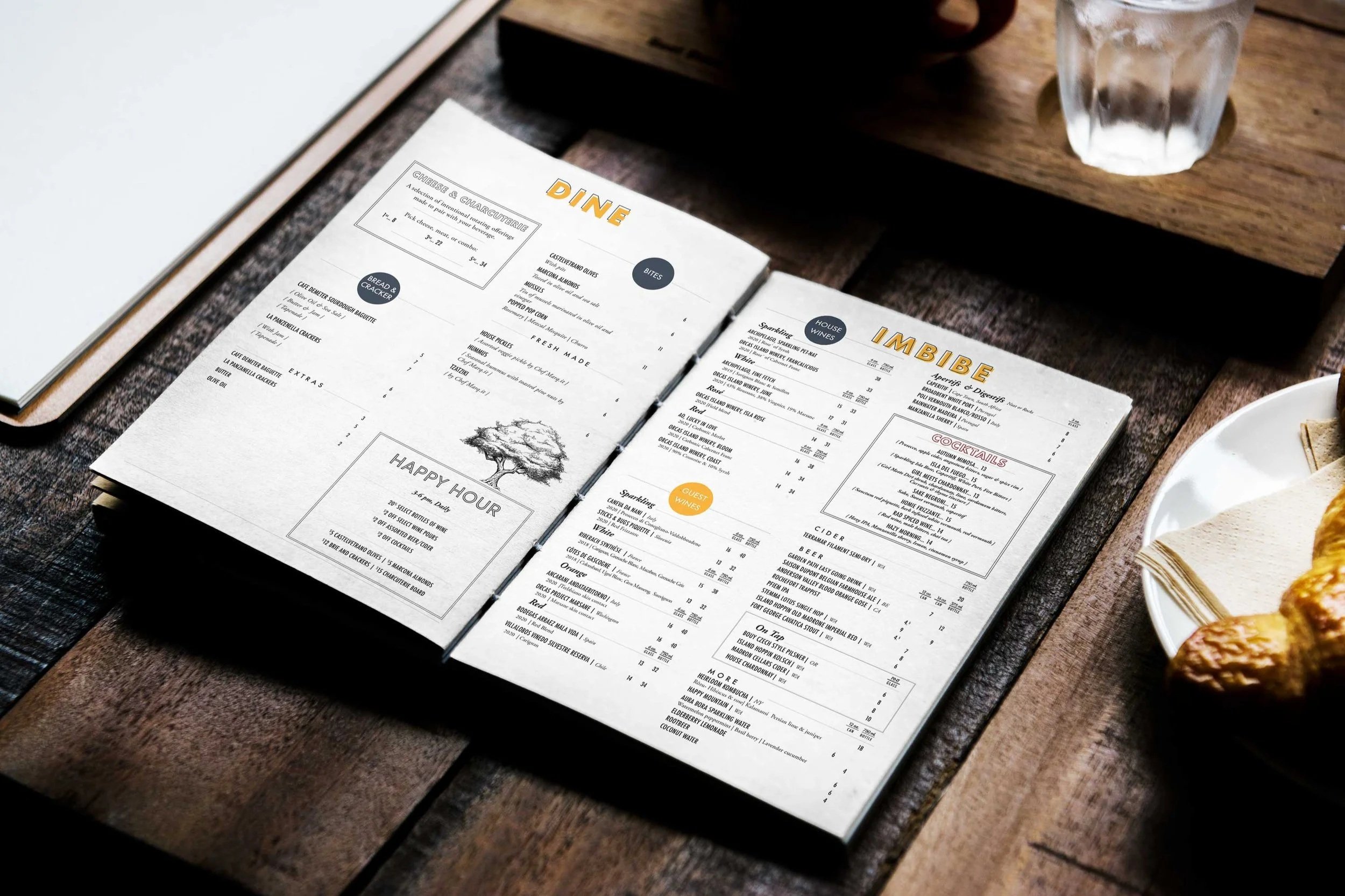 Restaurant menu open on a wooden table, featuring sections for dine, imbibe, and happy hour, with drinks and food options listed.
