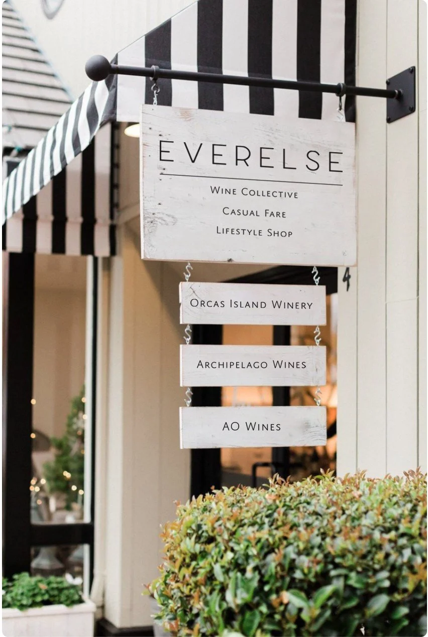 A white wooden sign hanging outside a shop called Everlse, showcasing that it is a wine collective, casual fare restaurant, and lifestyle shop. The sign also lists three wine vendors: Orcas Island Winery, Archipelago Wines, and AO Wines.