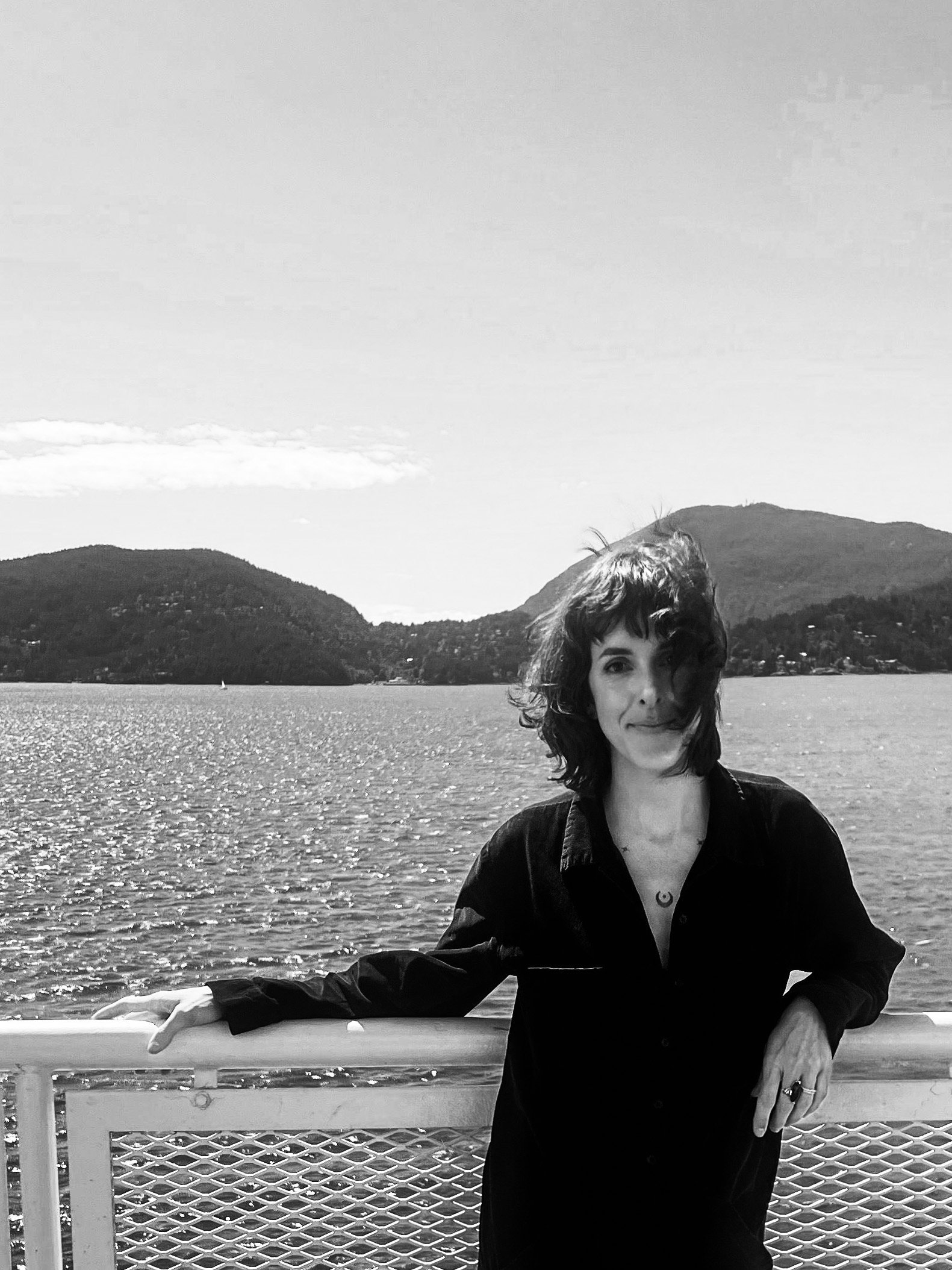 A woman with shoulder-length hair standing on a boat or ferry, leaning on a railing with a large lake and mountains in the background, black and white photo.